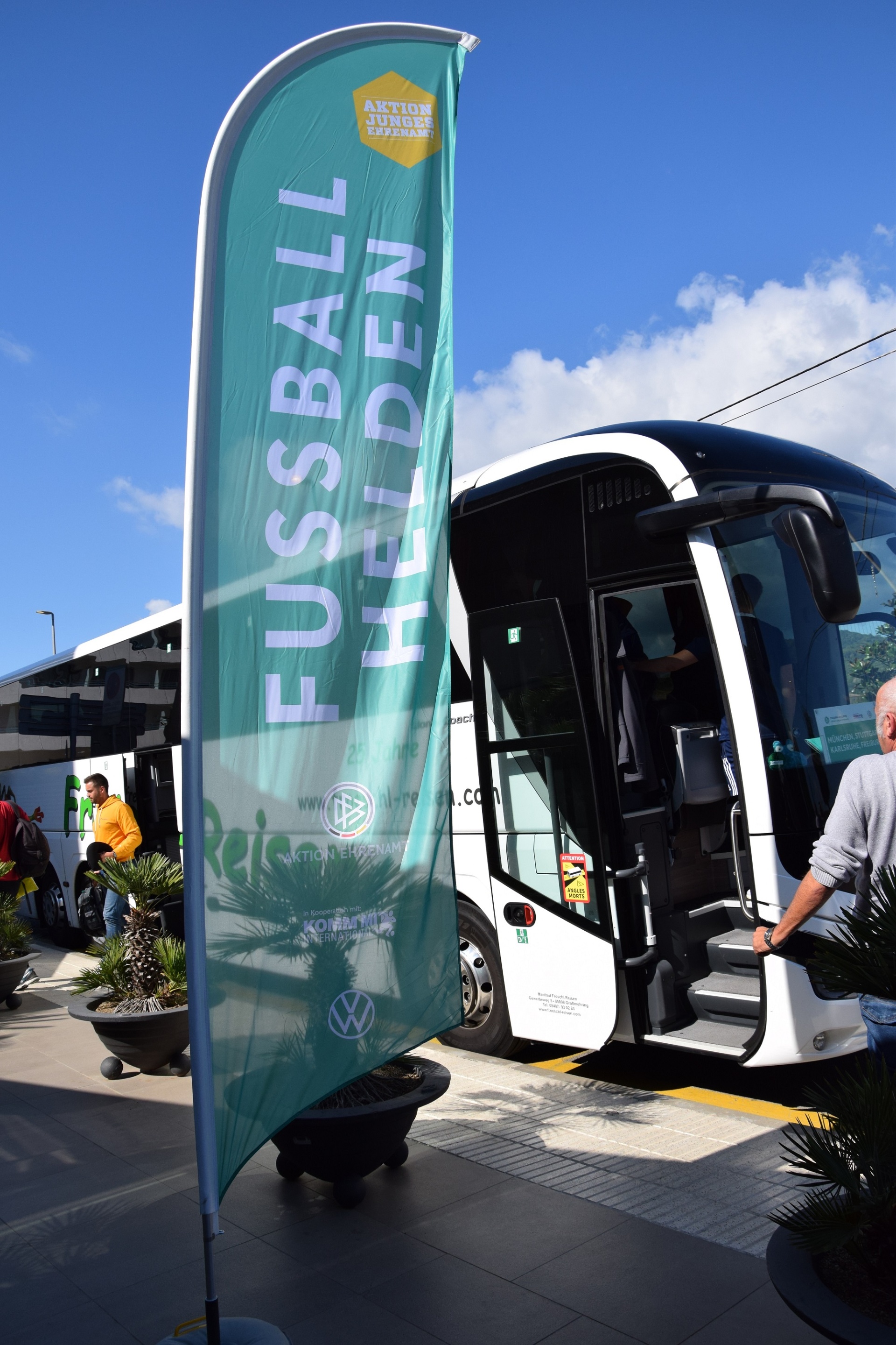 [{"clang_id":1,"value":"Weißer Reisebus mit geöffneter Tür. Links daneben eine grüne Fahne mit weißem Schriftzug \"FUSSBALL HELDEN\". Im Hintergrund Personen und Pflanzen."}]