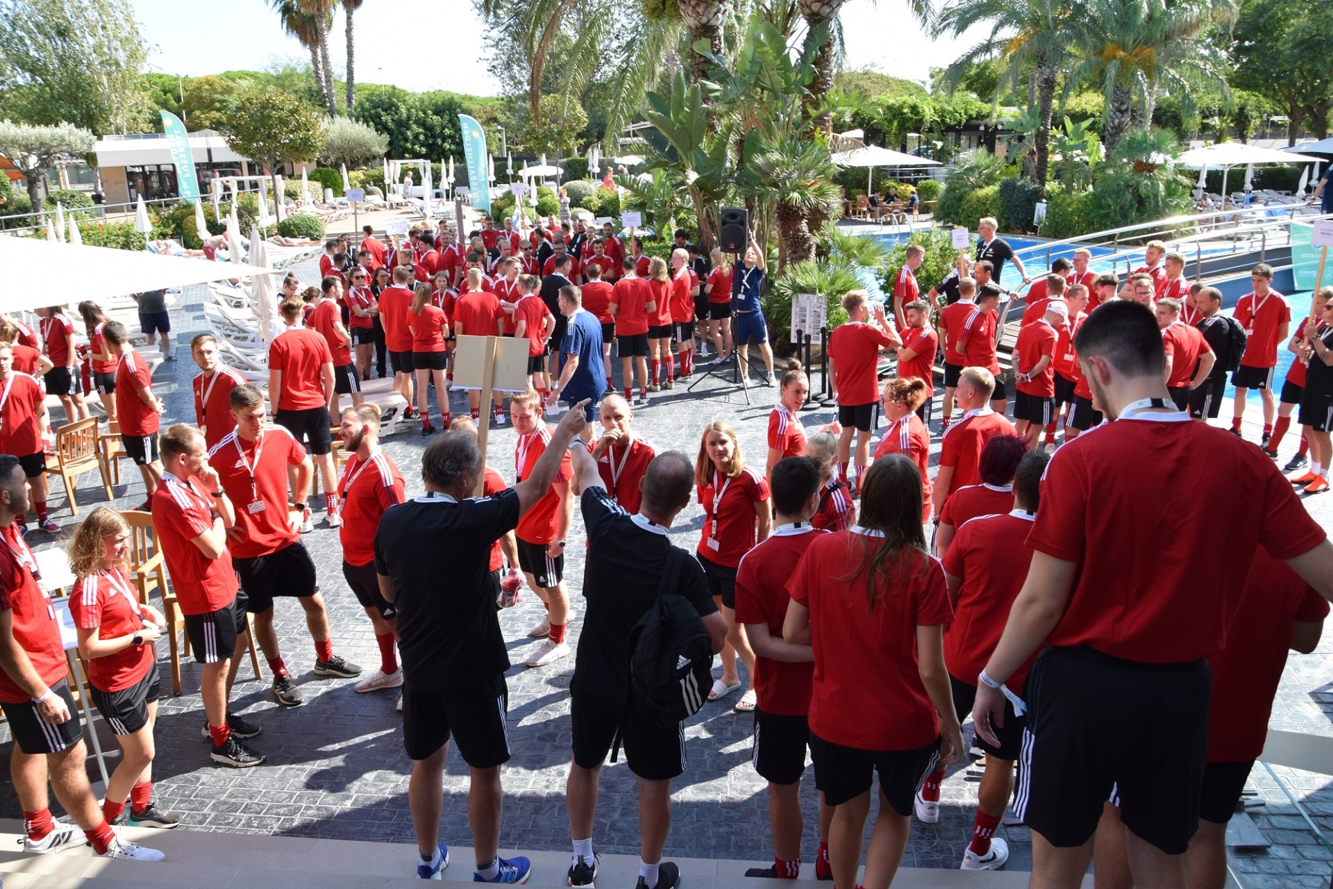 [{"clang_id":1,"value":"Große Gruppe Jugendlicher in roten Trainingsanzügen um einen Trainer herum, der Anweisungen gibt. Im Hintergrund Palmen und ein Schwimmbad."}]