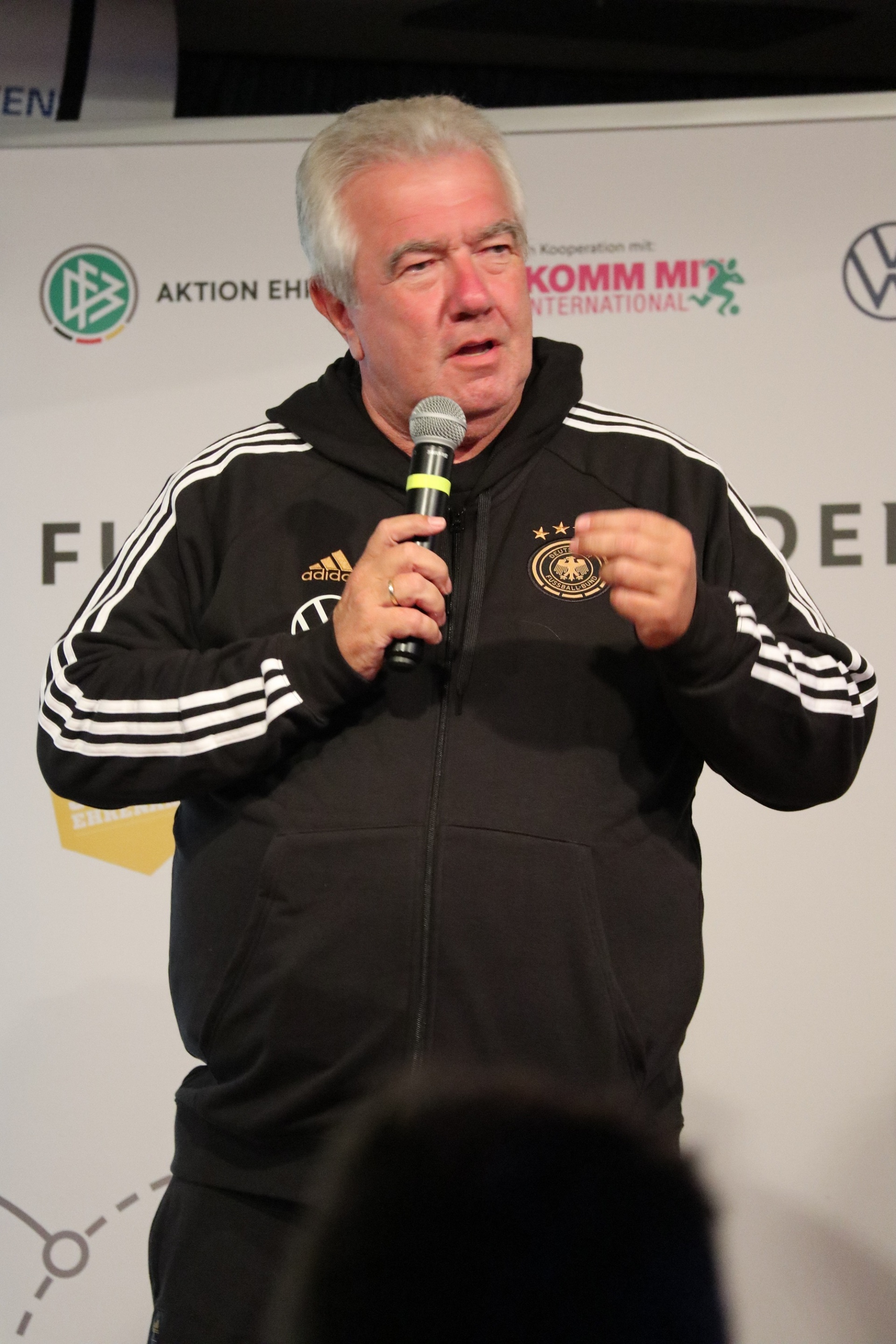 [{"clang_id":1,"value":"Mann mit weißen Haaren spricht in Mikrofon. Er trägt eine schwarze Trainingsjacke mit Adidas-Streifen und dem deutschen Fußballverband-Emblem. Hintergrund: Werbebanner."}]