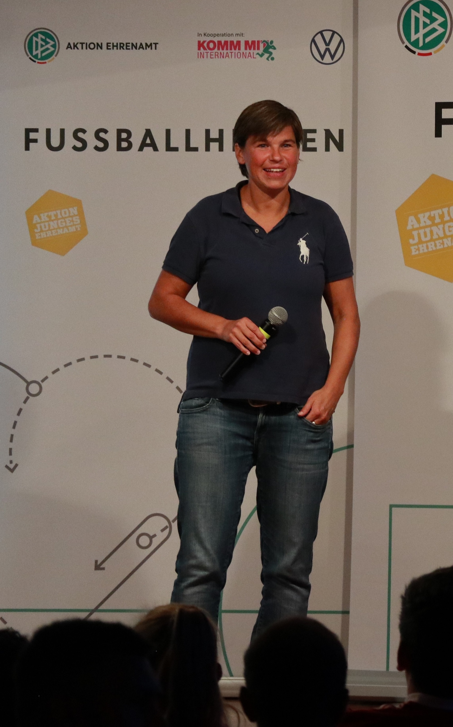 [{"clang_id":1,"value":"Frau mit Mikrofon steht auf einer Bühne vor einem Banner mit Fussballmotiv und Logos. Im Publikum sind Köpfe zu sehen."}]