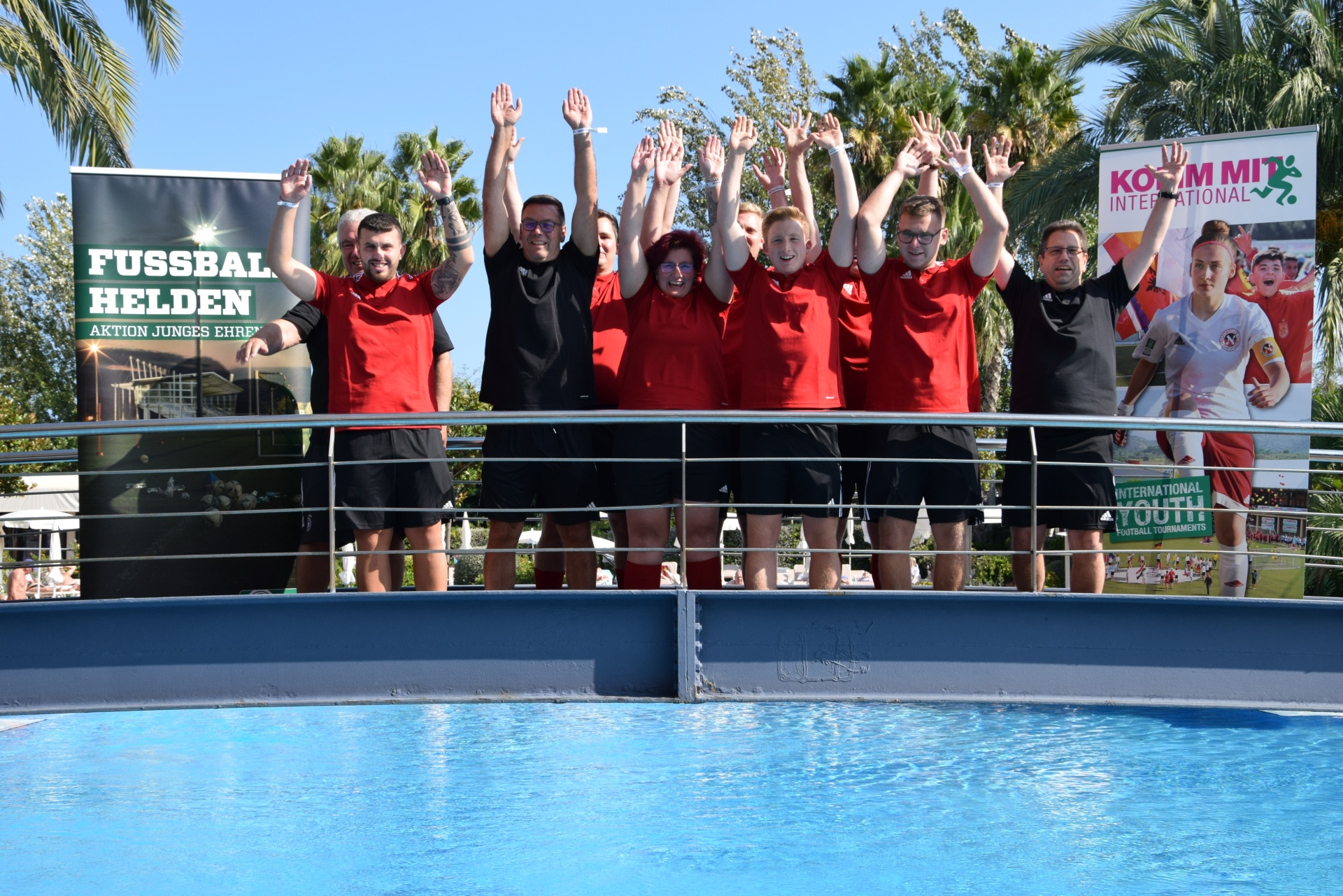 [{"clang_id":1,"value":"Gruppe von Personen mit hoch erhobenen Armen am Rand eines Swimmingpools. Im Hintergrund Banner mit Fußball-Motiv und Palmen."}]