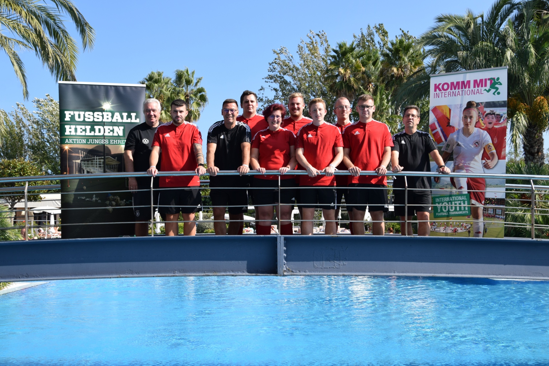 [{"clang_id":1,"value":"Gruppe von neun Personen steht lächelnd vor einem Pool und Werbebannern. Sie tragen rote und schwarze Sportkleidung. Palmen im Hintergrund."}]