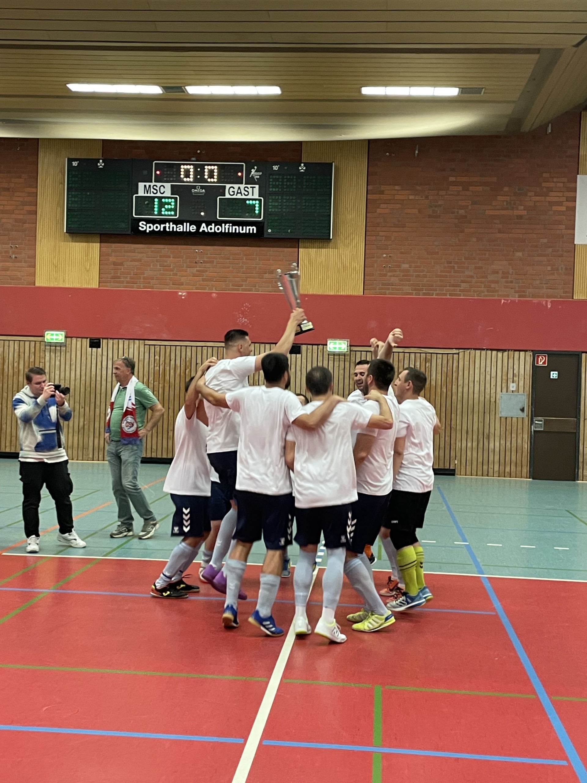 [{"clang_id":1,"value":"Handballteam feiert Sieg in Sporthalle. Spieler im Kreis, halten Pokal hoch. Scoreboard im Hintergrund zeigt Ergebnis. Zuschauer am Rand."}]