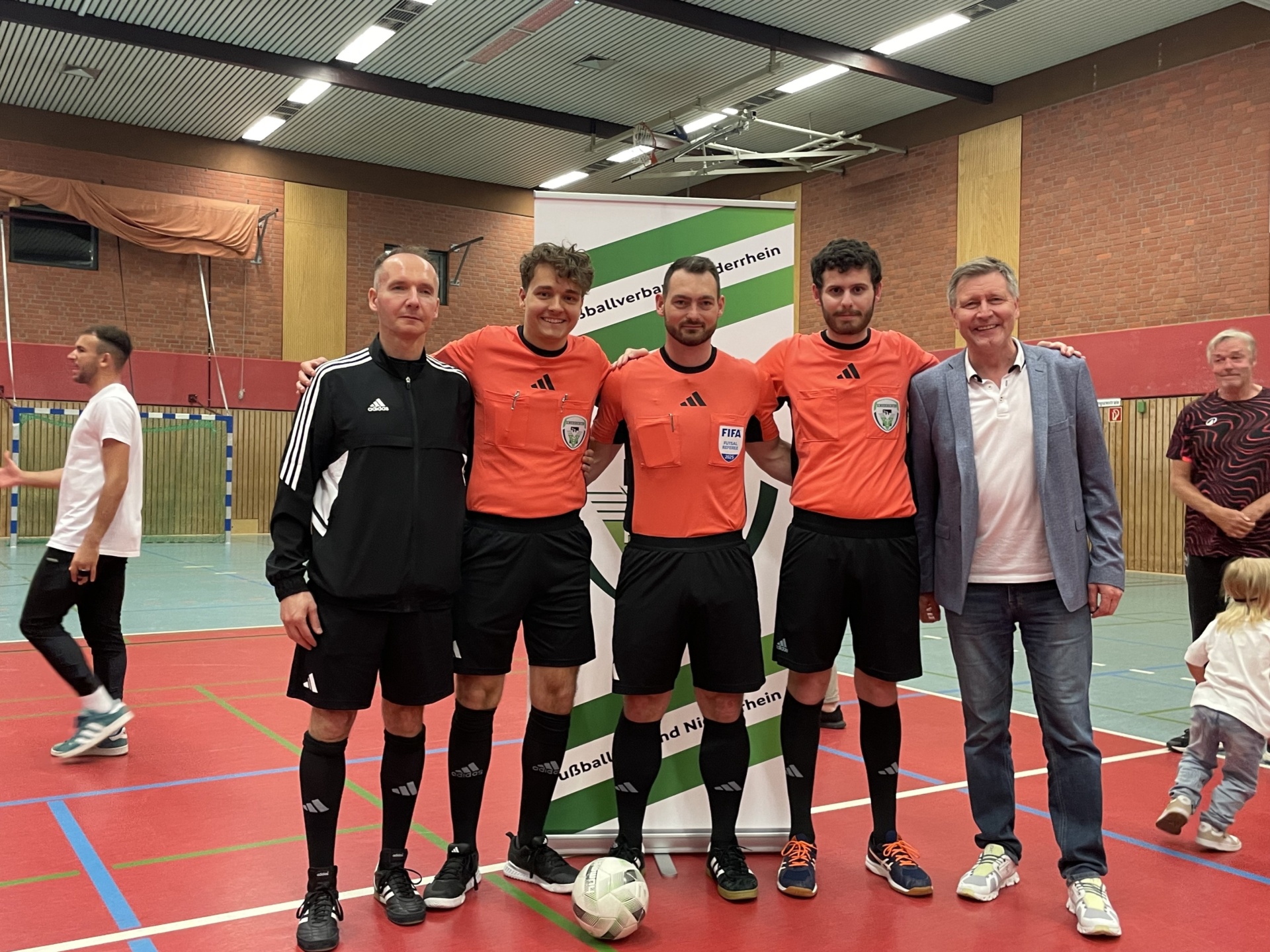 [{"clang_id":1,"value":"Fünf Männer stehen in einer Sporthalle. Drei in rot-schwarzen Schiedrichteroutfits, einer in Schwarz-Weiß, ein weiterer in dunklem Anzug. Ein Fußball liegt vor ihnen. Im Hintergrund eine Bannerwerbung."}]