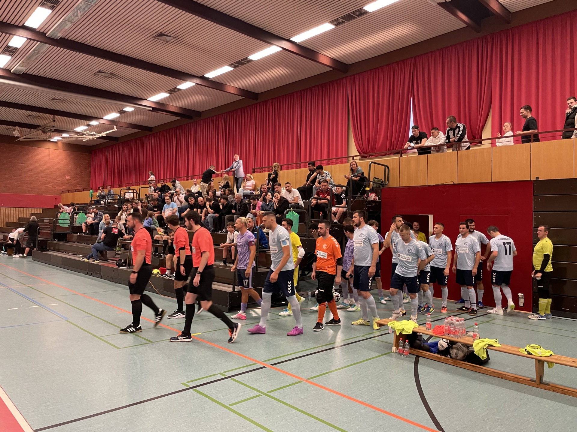 [{"clang_id":1,"value":"Handballspiel in Sporthalle. Zwei Teams in Sportkleidung auf dem Feld, Schiedsrichter, Zuschauer auf Tribüne mit rotem Vorhang im Hintergrund."}]