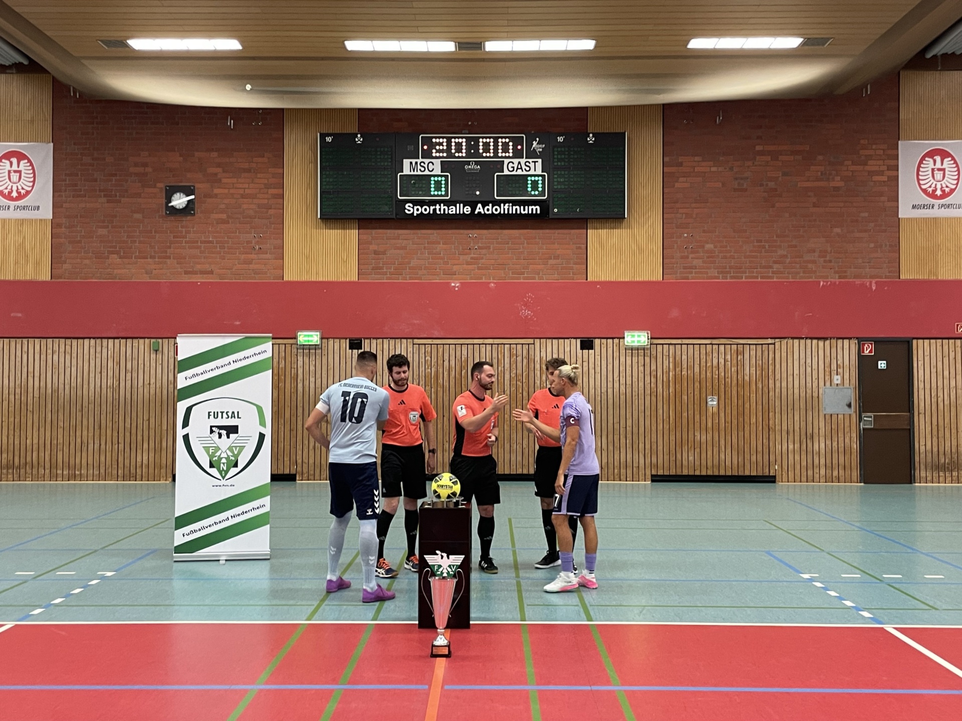 [{"clang_id":1,"value":"Sporthalle mit Futsal-Spielfeld. Gruppe Personen, darunter Schiedsrichter, steht um einen Pokal. Digitale Anzeigetafel zeigt Spielzeit."}]