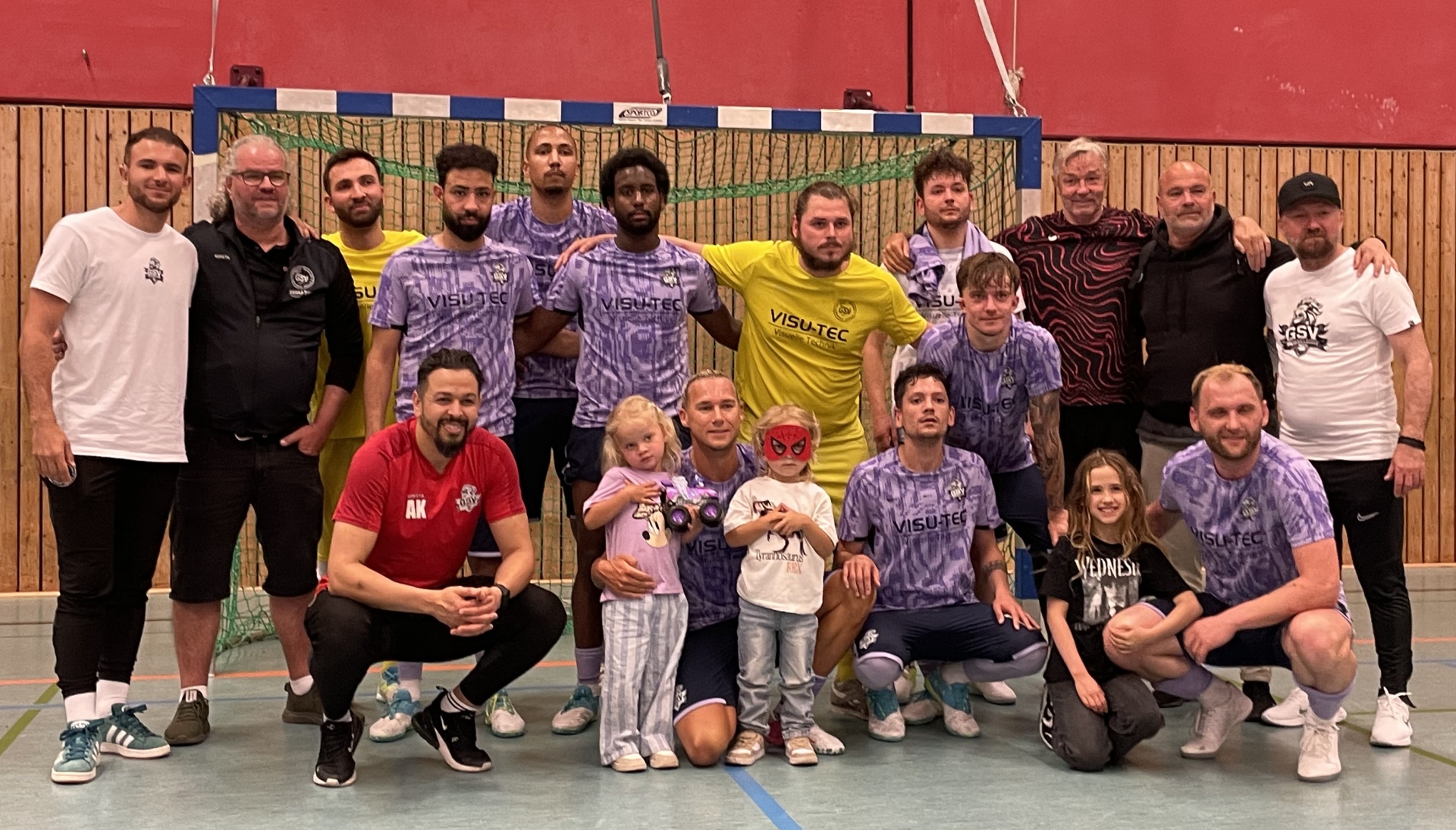 [{"clang_id":1,"value":"Fussballmannschaft mit Kindern und Betreuern posiert vor einem Tor in einer Sporthalle. Alle tragen Sportkleidung, meist in Lila- und Schwarztönen."}]