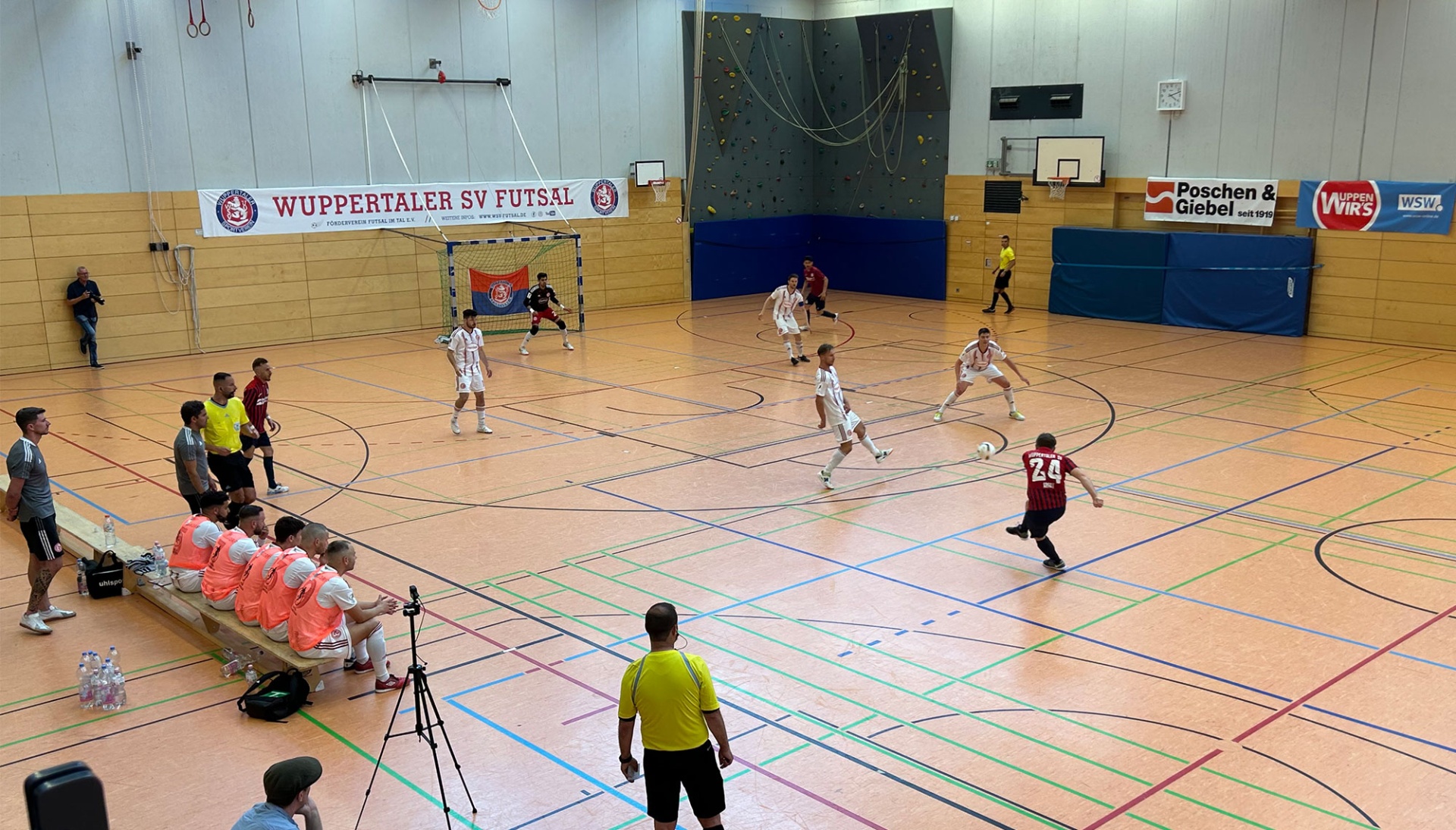 [{"clang_id":1,"value":"Futsal-Spiel in Sporthalle. Zwei Teams in roten und weißen Trikots spielen auf bunt markiertem Boden. Zuschauer auf Banken, Schiedsrichter, Kamera-Ausrüstung. Banner \"Wuppertaler SV Futsal\"."}]
