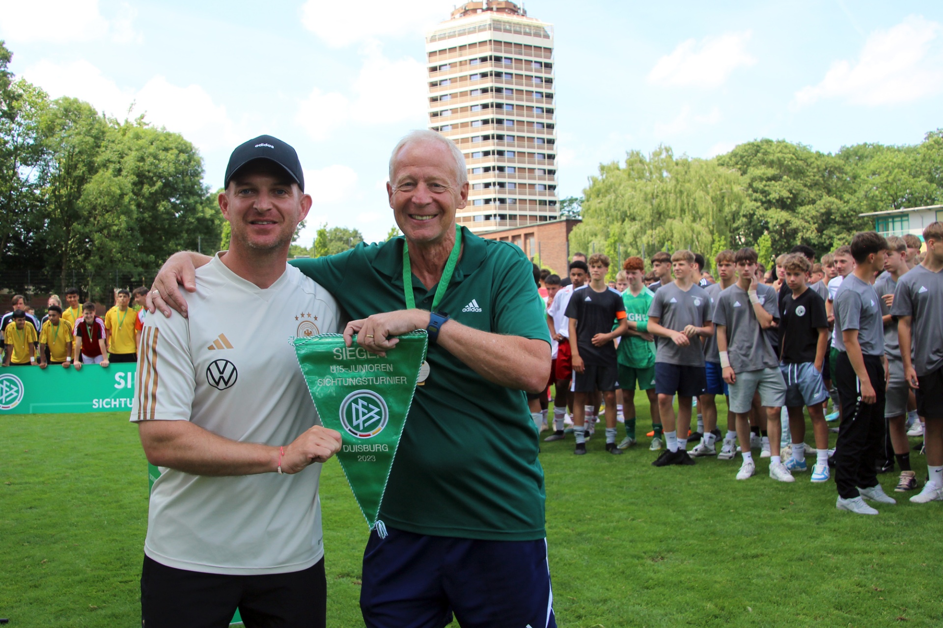 [{"clang_id":1,"value":"Zwei Männer stehen auf einem Fußballfeld vor einer Gruppe Jugendlicher und einem Hochhaus. Der Mann links trägt Kappe und Trainingsanzug, der rechte einen Pullover mit Wappen. Beide halten eine blau-weiße Flagge."}]
