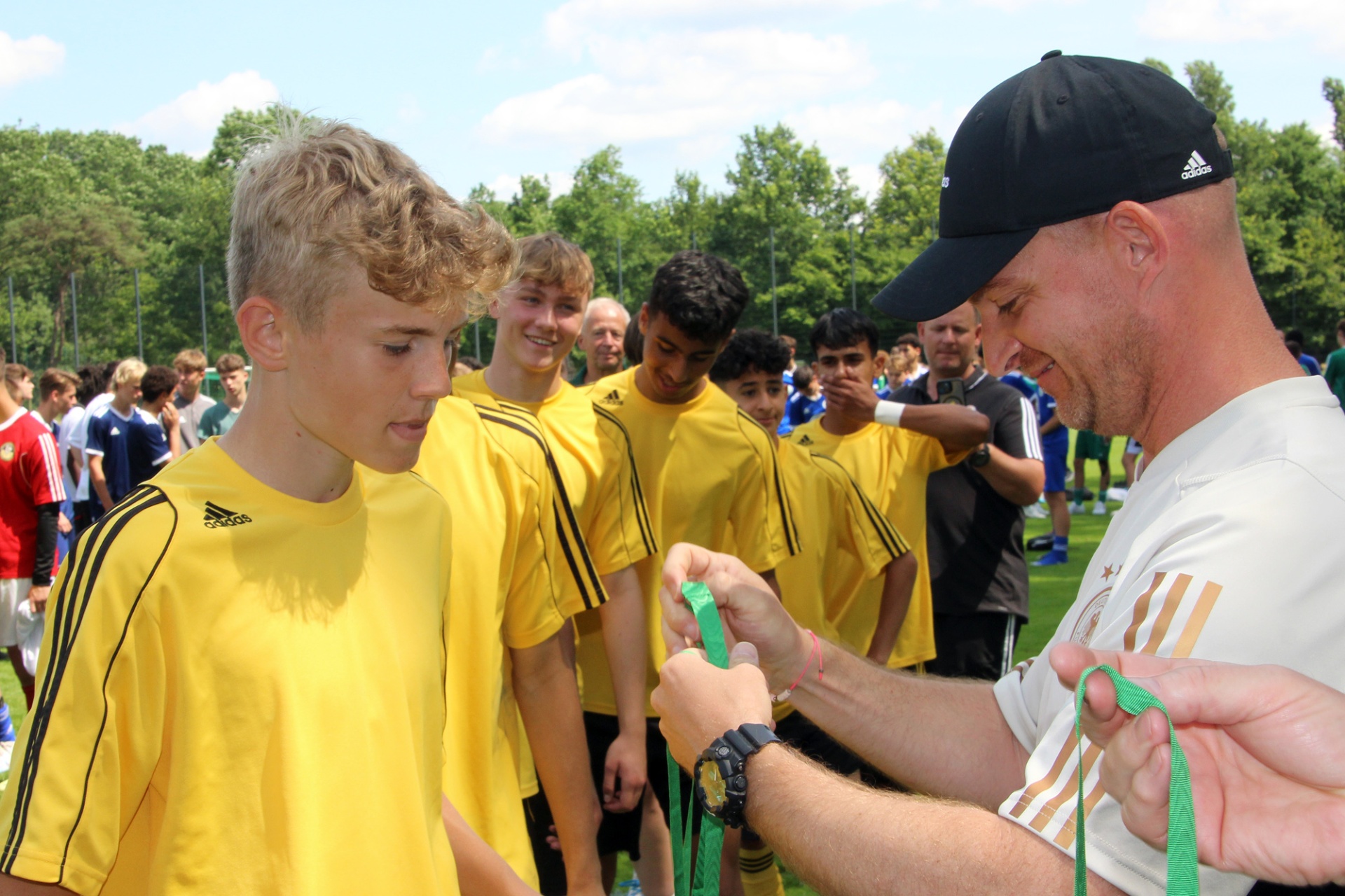 [{"clang_id":1,"value":"Fußballspieler in gelben Trikots und ein Trainer mit schwarzer Kappe. Der Trainer befestigt einem Spieler ein grünes Band an der Sportausrüstung."}]