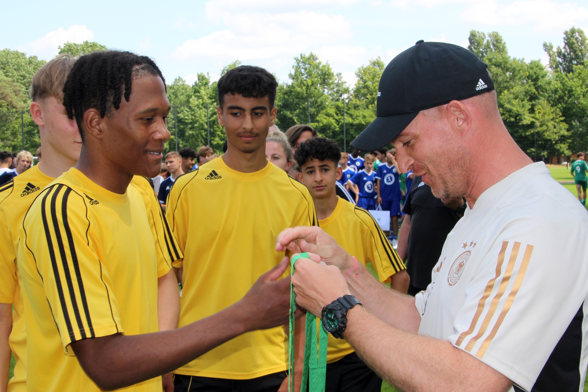 [{"clang_id":1,"value":"Fußballtrainer bindet einem Jugendlichen einen grünen Armverband. Im Hintergrund weitere Fußballspieler mit gelben Trikots und Bäume."}]