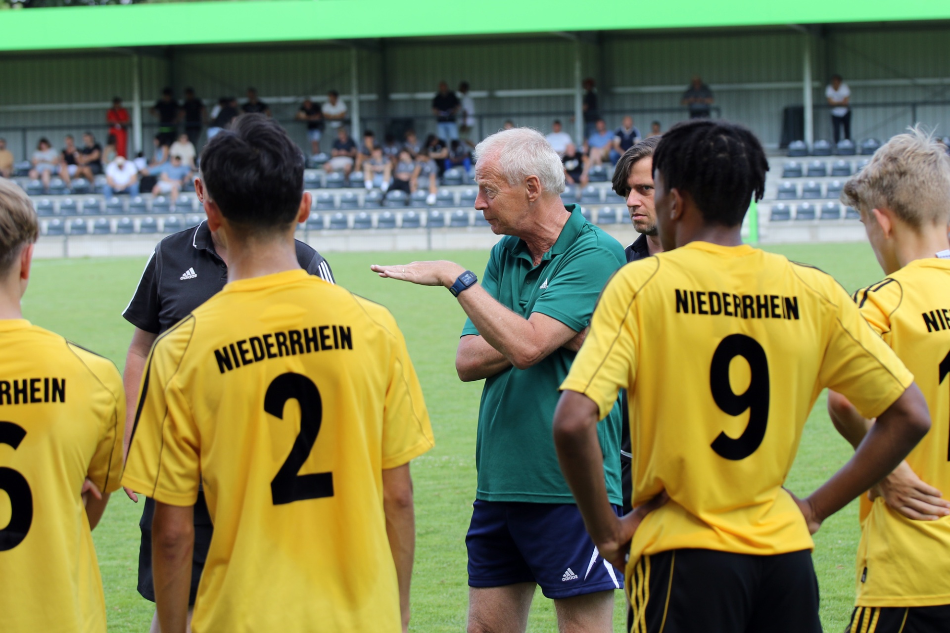 [{"clang_id":1,"value":"Fußballtrainer mit Pfeife bespricht Taktik mit vier Spielern in gelben Trikots mit Aufschrift \"NIEDERRHEIN\" auf dem Fußballfeld. Im Hintergrund Zuschauer."}]