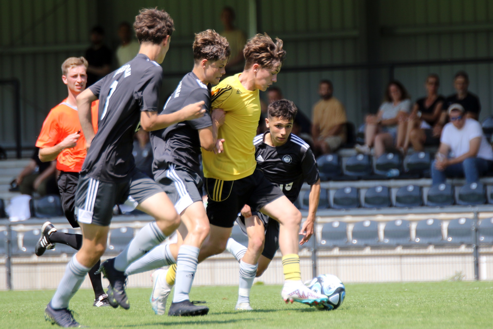 [{"clang_id":1,"value":"Fußballspiel: Vier Spieler – zwei in Schwarz, einer in Gelb und einer in Orange – kämpfen um den Ball auf einem grünen Rasenfeld. Im Hintergrund Zuschauerreihen."}]