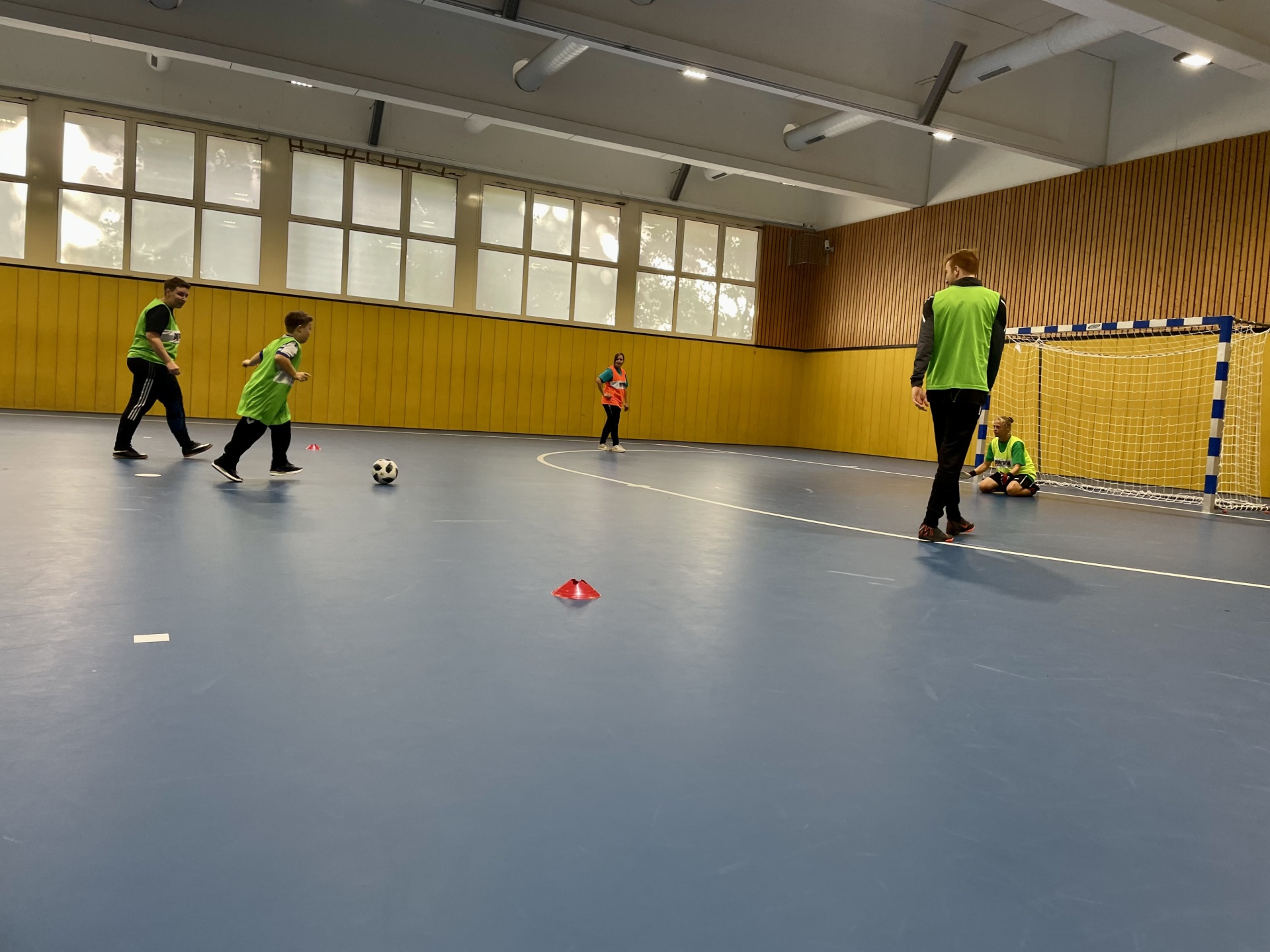 [{"clang_id":1,"value":"Sporthalle mit blauem Boden. Kinder und Trainer beim Fußballtraining. Im Hintergrund eine gelbe Wand mit Fenstern und ein Handballtor."}]