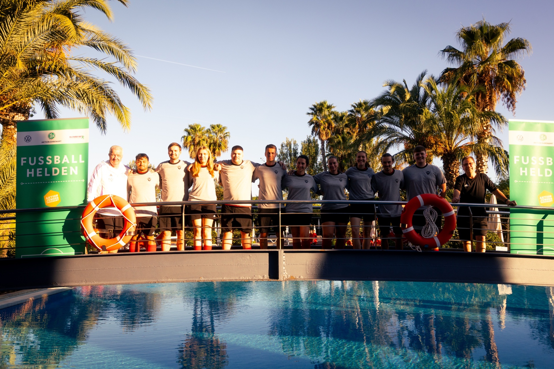 [{"clang_id":1,"value":"Gruppe von Personen vor Pool und Werbebannern mit Schriftzug \"Fussball Helden\". Palmen im Hintergrund, sonniger Tag. Rettungsringe an der Poolumrandung."}]