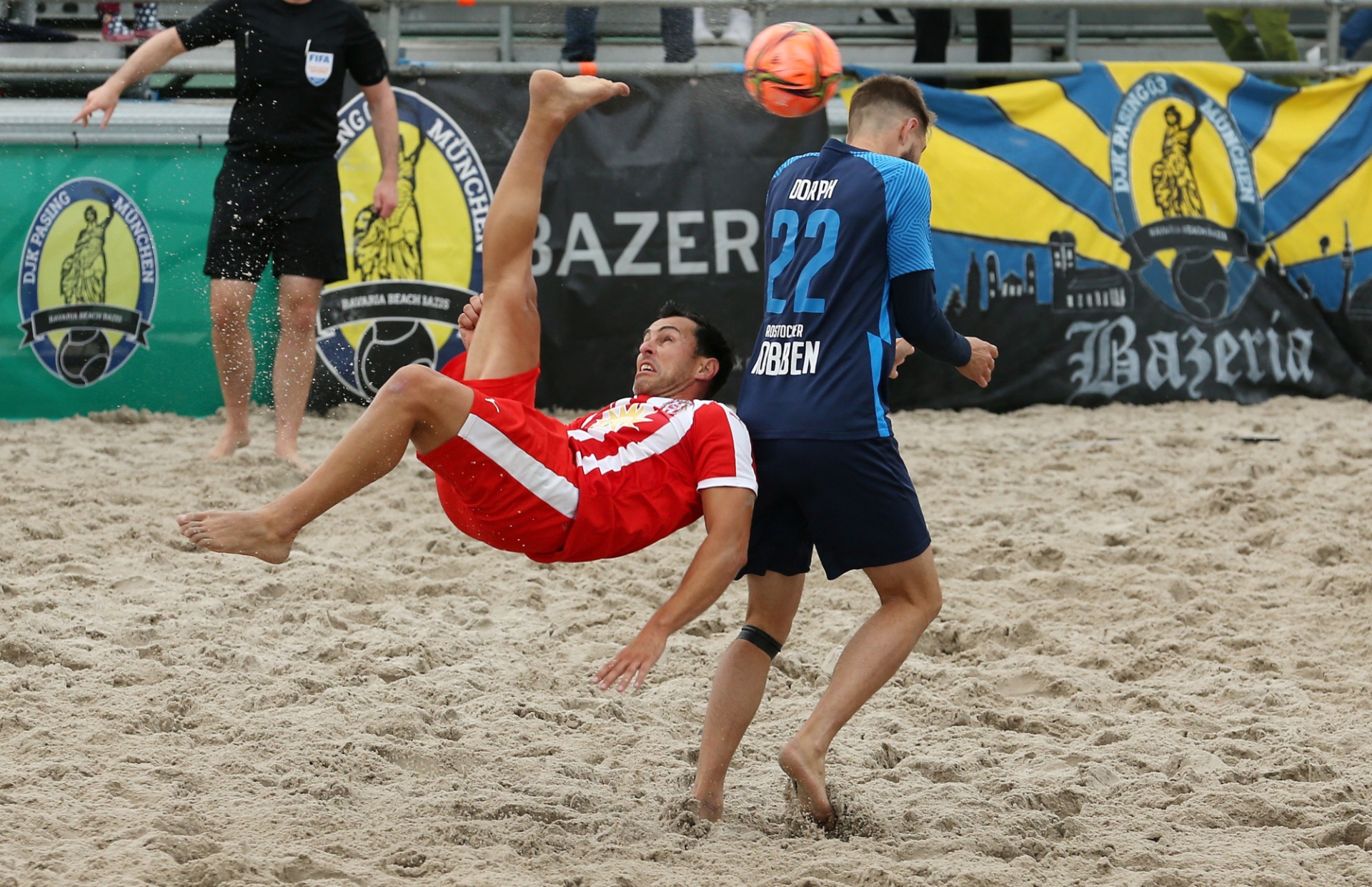 [{"clang_id":1,"value":"Zwei Beachvolleyballspieler im Sand. Ein Spieler in Rot vollführt einen Rückwärts-Saltospringer, der andere in Blau versucht den Ball zu spielen. Im Hintergrund weitere Spieler und Werbebanner."}]