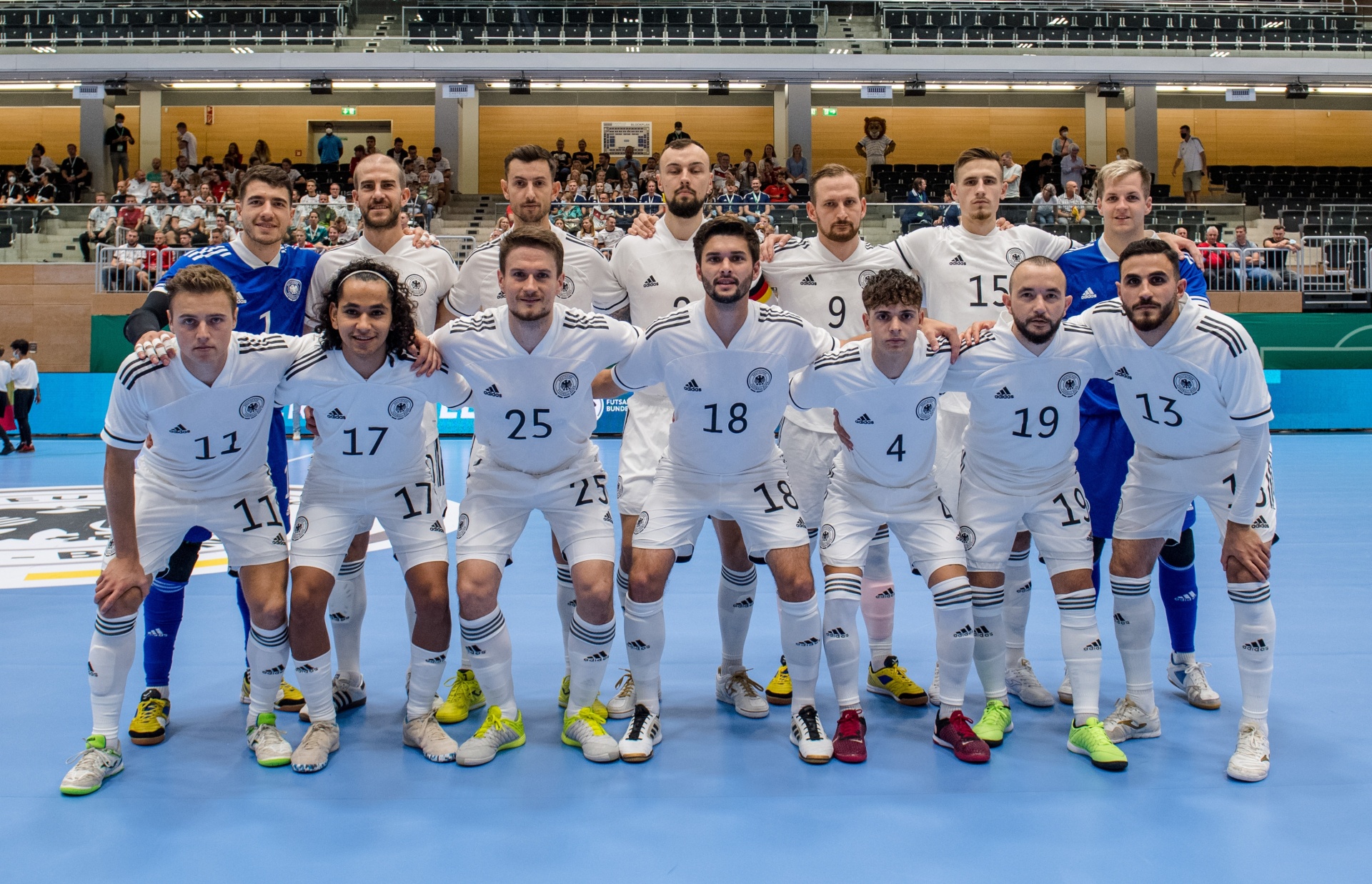 [{"clang_id":1,"value":"Futsal-Mannschaft in weißen Trikots mit Nummern (4-25) und blauen Shorts. Spieler knien und stehen auf blauem Hallenboden vor Zuschauerrängen."}]