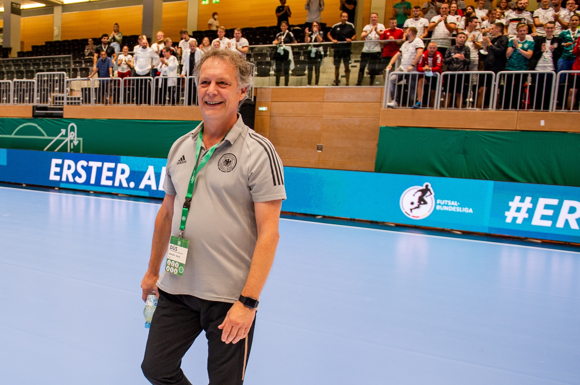 [{"clang_id":1,"value":"Mann mit Polohemd und Lanyard steht lächelnd auf einer blauen Futsal-Halle. Im Hintergrund Zuschauerreihen. Werbebanner sind sichtbar."}]