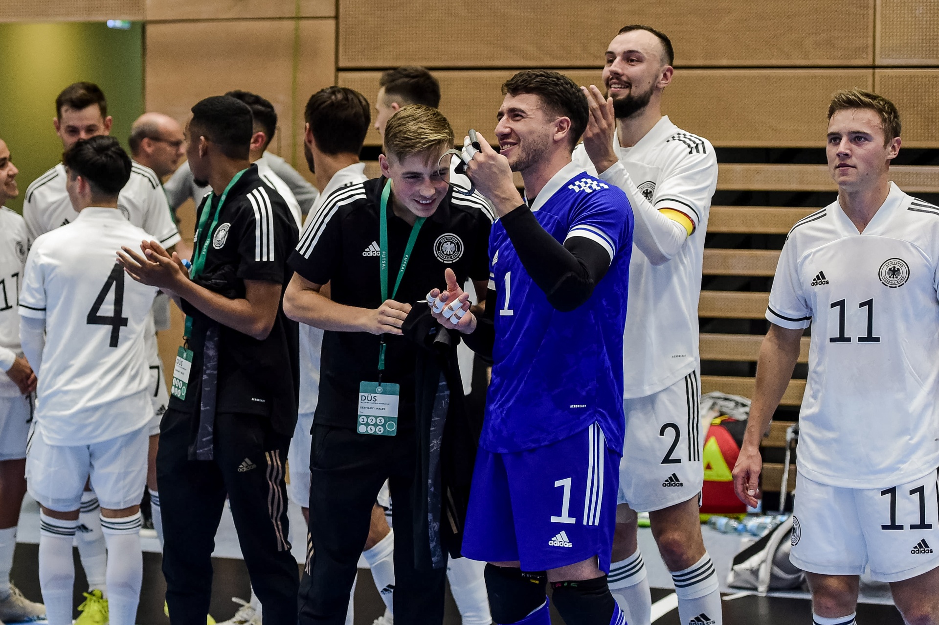 [{"clang_id":1,"value":"Futsal-Spieler in weißen und blauen Trikots feiern oder begrüßen sich in einem Gang. Ein Mann mit schwarzem Shirt und Lanyard steht im Mittelpunkt."}]