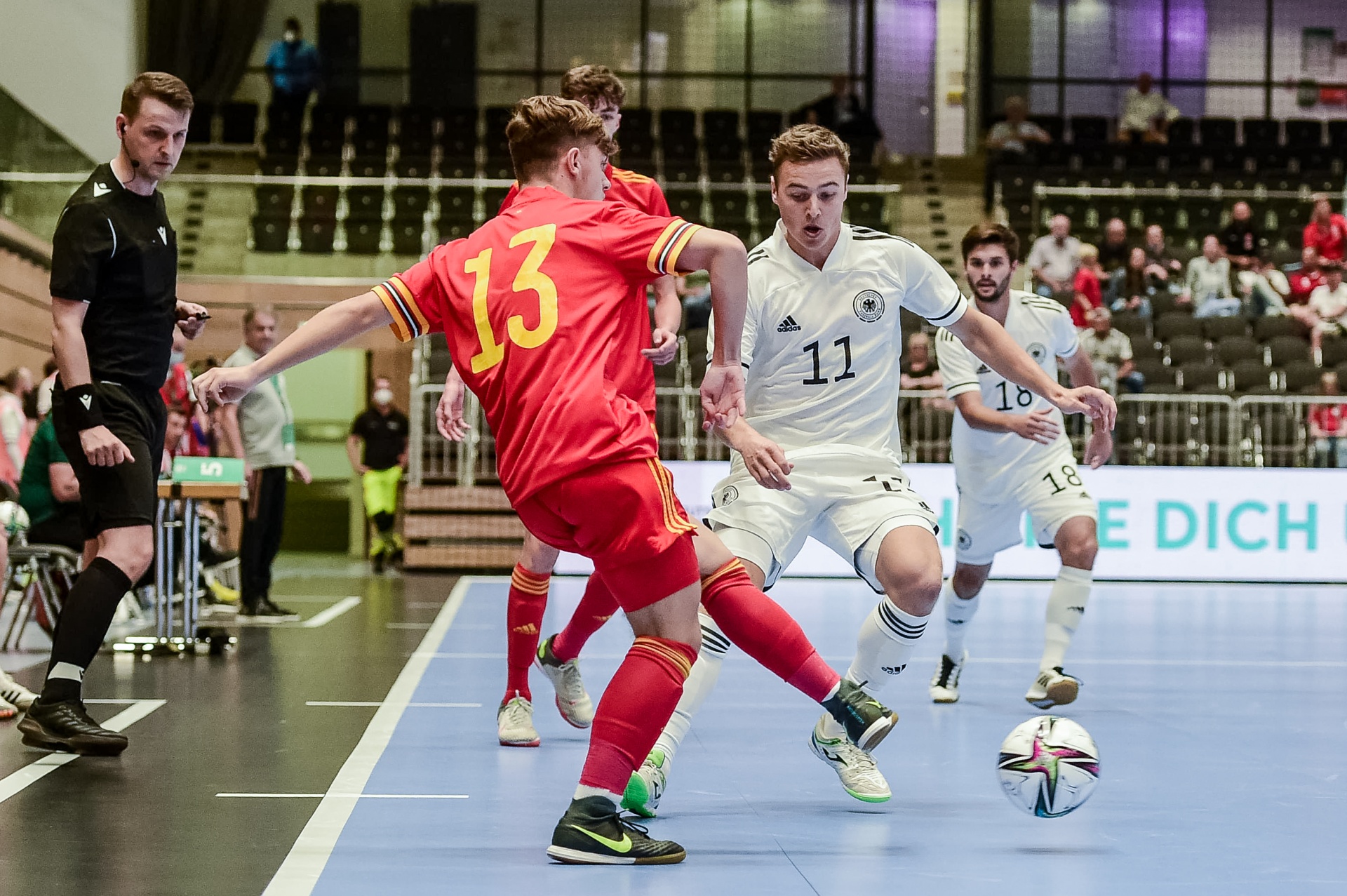 [{"clang_id":1,"value":"Futsalspiel: Spieler mit Trikotnummer 11 (Deutschland) dribbelt den Ball, wird von Spieler mit Nummer 13 (Belgien) gefordert. Schiedsrichter und Zuschauer im Hintergrund."}]