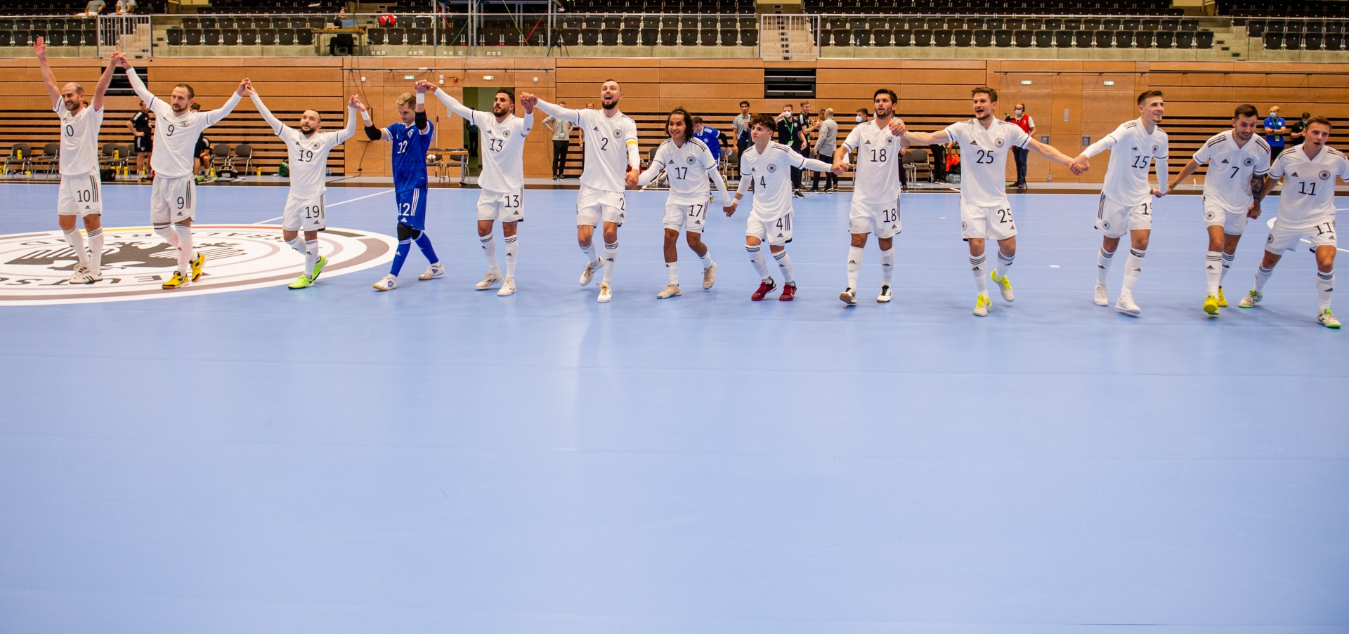 [{"clang_id":1,"value":"Handballmannschaft in blauen und weißen Trikots steht auf blauem Spielfeld mit erhobenen Händen. Im Hintergrund leere Zuschauertribünen."}]