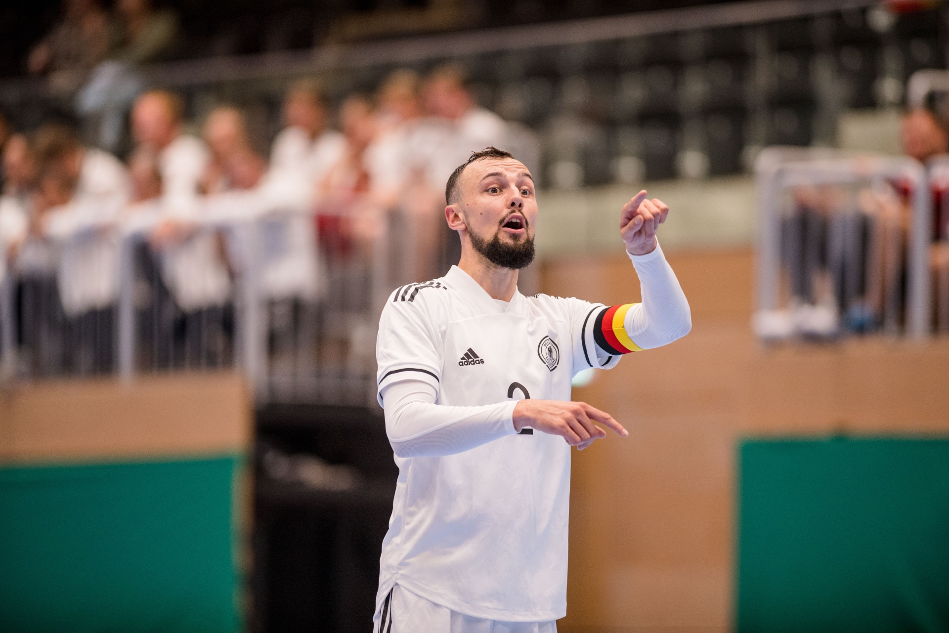 [{"clang_id":1,"value":"Handballspieler mit Bart im weißen Trikot der deutschen Nationalmannschaft, Arme gehoben, vor verschwommener Zuschauerkulisse in Sporthalle."}]