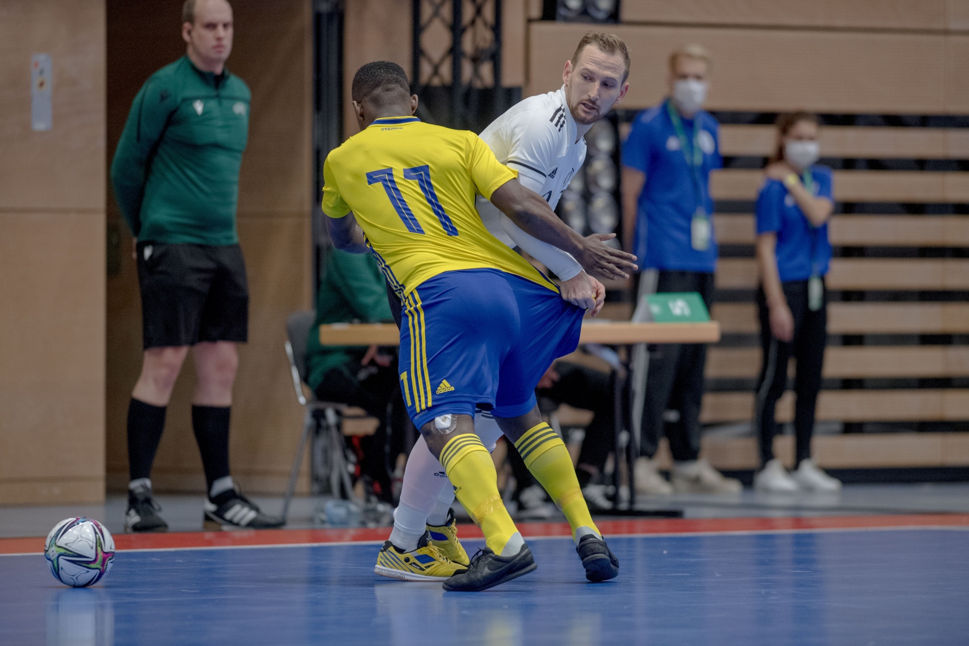 [{"clang_id":1,"value":"Zwei Futsalspieler in blauen und gelben bzw. weißen Trikots im Zweikampf. Im Hintergrund Zuschauer und ein Schiedsrichter. Ein Fußball liegt am Boden."}]