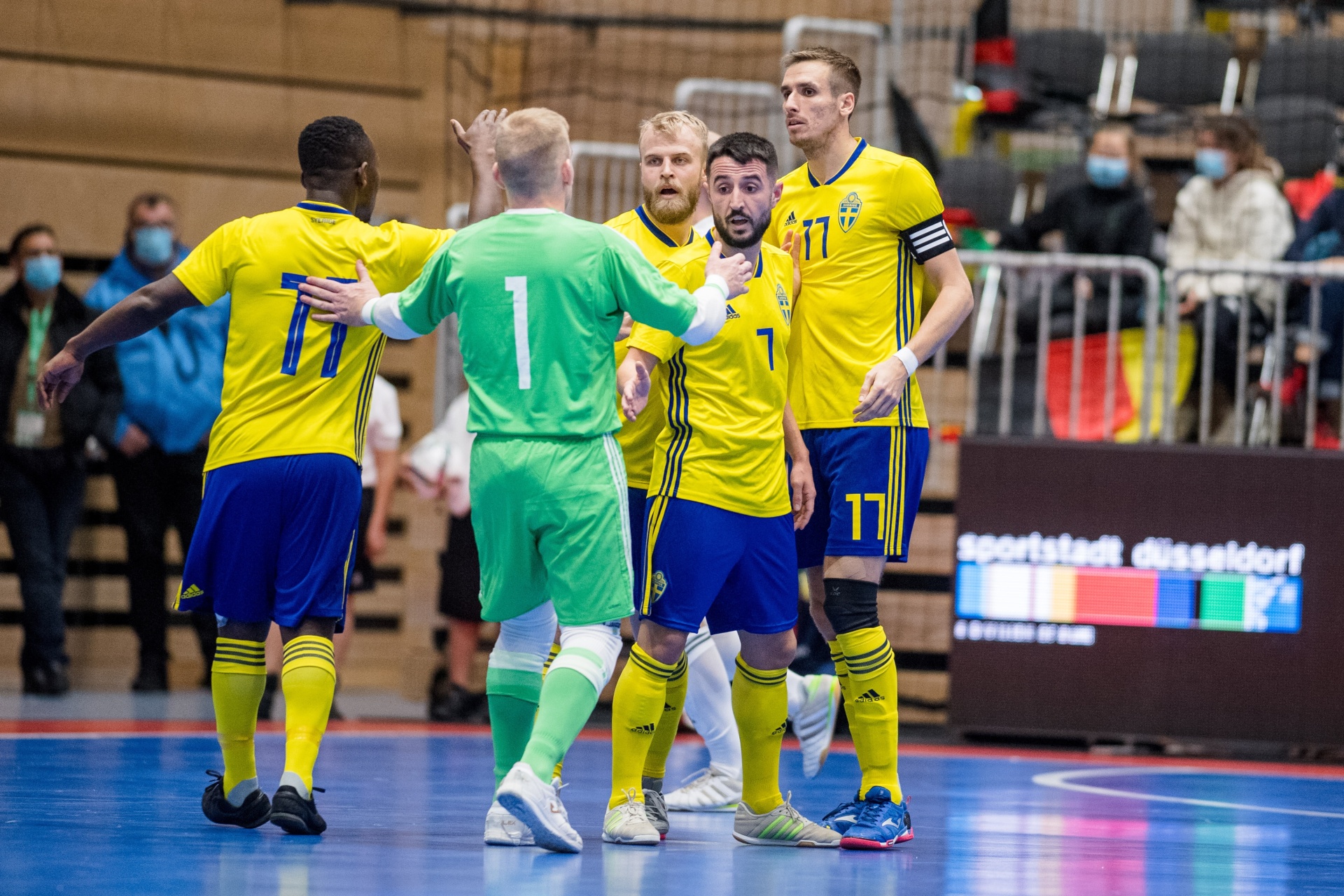 [{"clang_id":1,"value":"Futsal-Spieler in blauen und gelben Trikots feiern auf dem Hallenfußballfeld. Im Hintergrund Zuschauer und das Logo \"Sportstadt Düsseldorf\"."}]