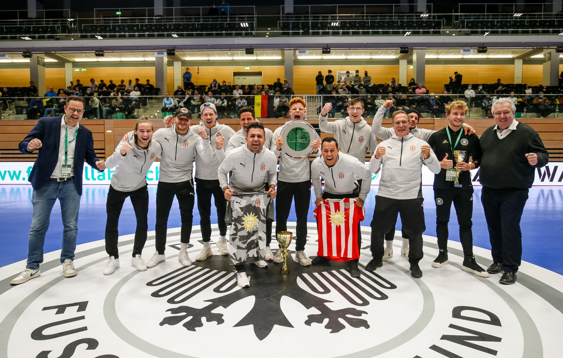 [{"clang_id":1,"value":"Fußballmannschaft in hellgrauen Trainingsanzügen und rot-weißen Schals posieren mit Pokal auf einem Hallenboden mit Vereinslogo. Zwei Betreuer stehen an den Seiten."}]
