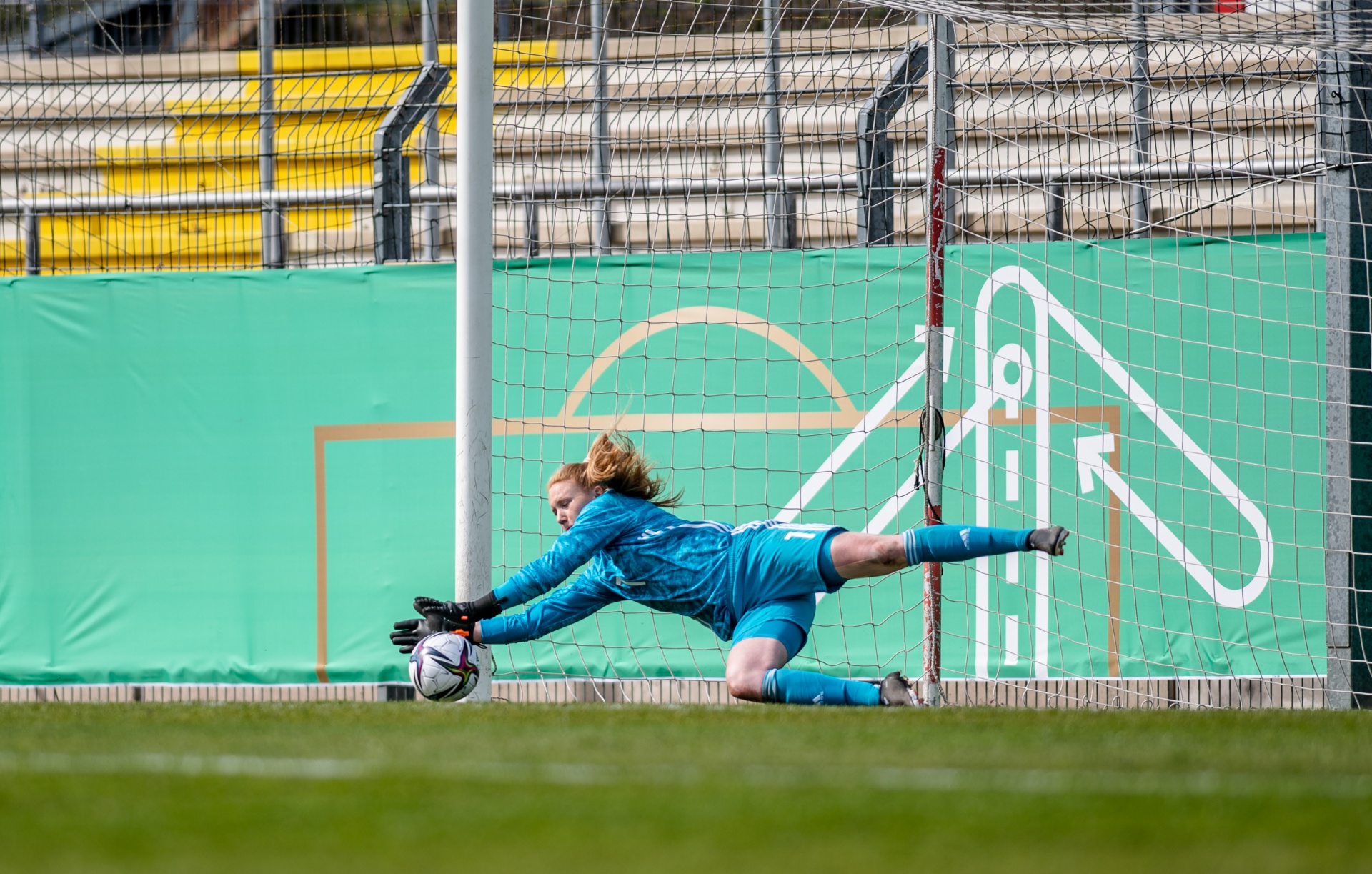 [{"clang_id":1,"value":"Fußballtorhüterin in blauer Ausrüstung streckt sich beim Fangen eines Balls im Spiel aus. Hintergrund: Stadiontribüne und grüner Zaun."}]