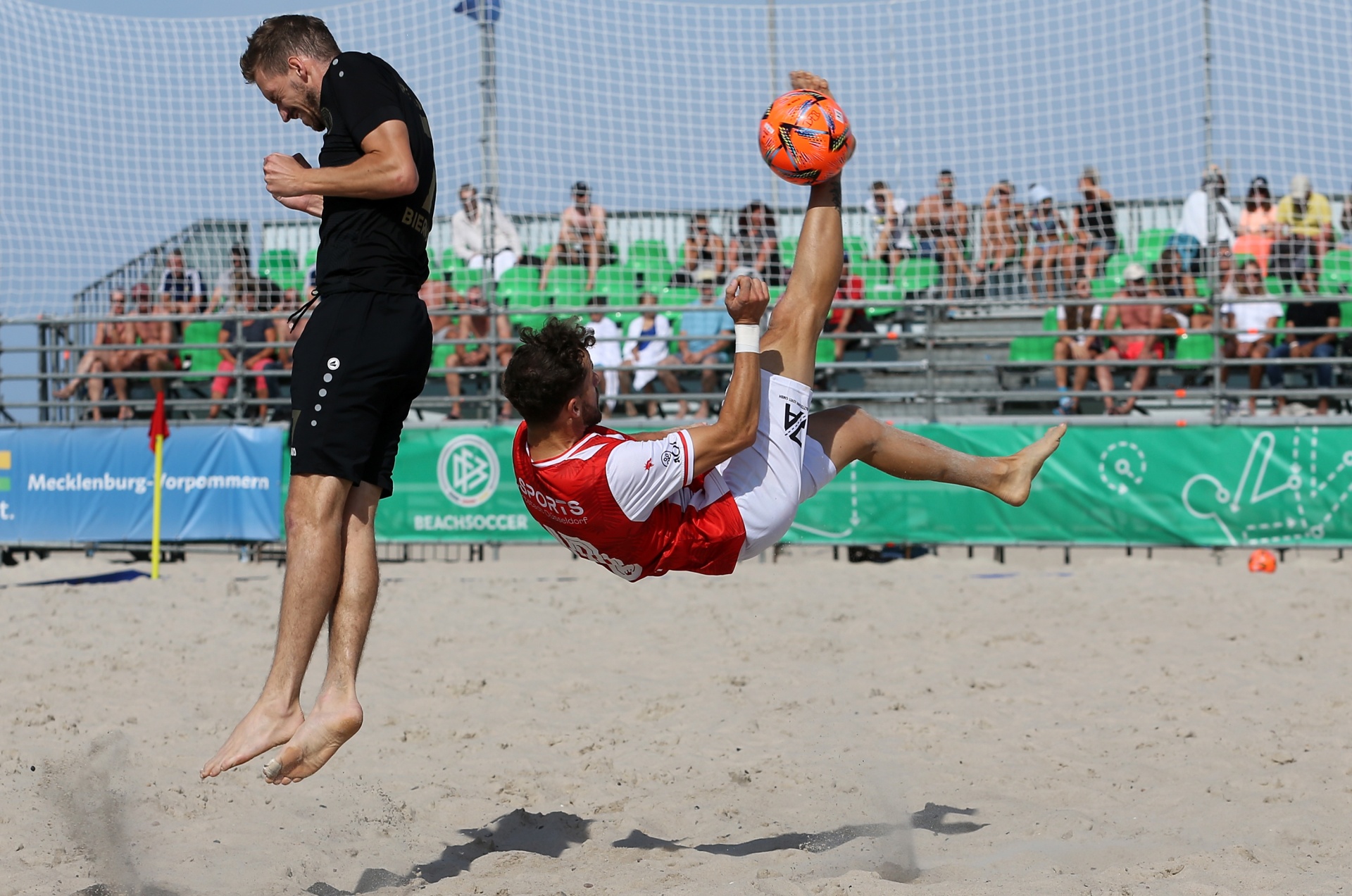 [{"clang_id":1,"value":"Zwei Beachsoccer-Spieler im Aktion. Ein Spieler springt, um den Ball mit der Ferse zu treten, während der andere den Ball verteidigt. Sandstrand, Zuschauerkulisse im Hintergrund."}]