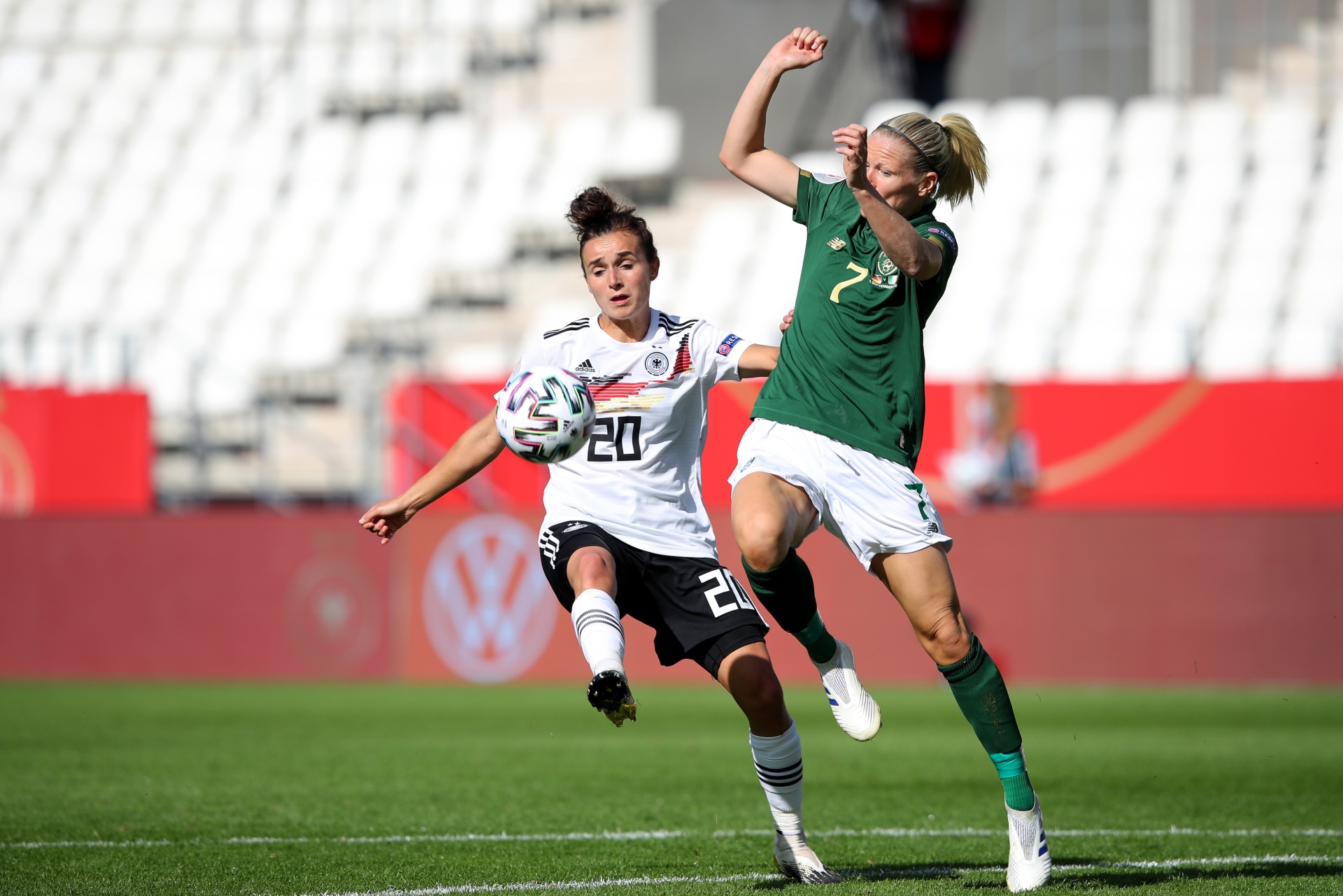 [{"clang_id":1,"value":"Zwei Fußballspielerinnen im Zweikampf auf grünem Rasen. Links eine Spielerin mit Nummer 20 im schwarz-weiß gestreiften Trikot, rechts eine Spielerin mit Nummer 7 im grünen Trikot. Im Hintergrund eine Zuschauertribüne."}]