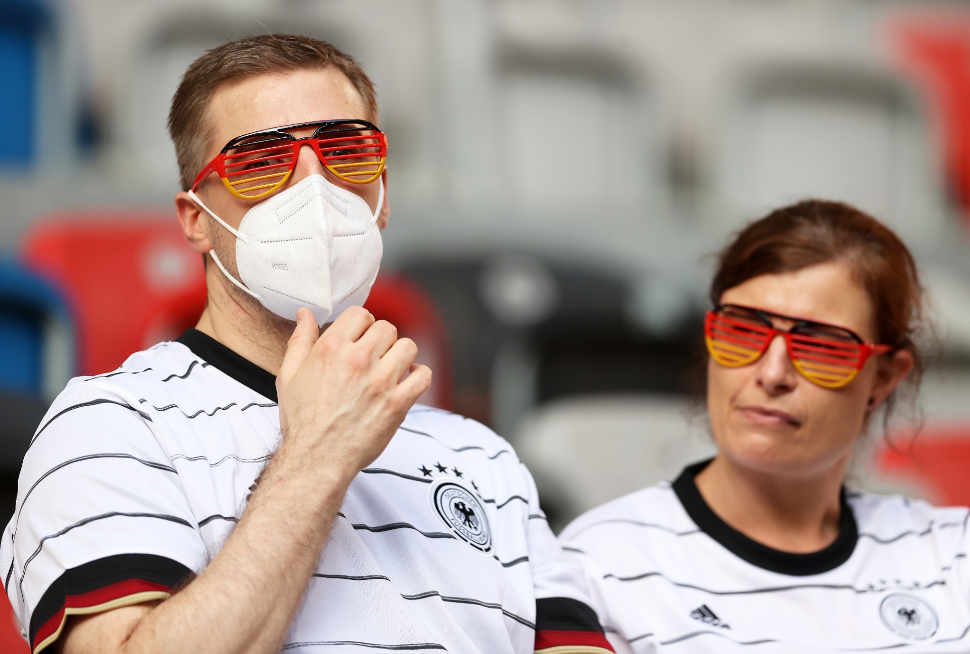 [{"clang_id":1,"value":"Zwei Fans mit Deutschlandtrikots und roter Sonnenbrille, der Mann trägt zusätzlich eine Maske. Hintergrund ist ein Stadion."}]