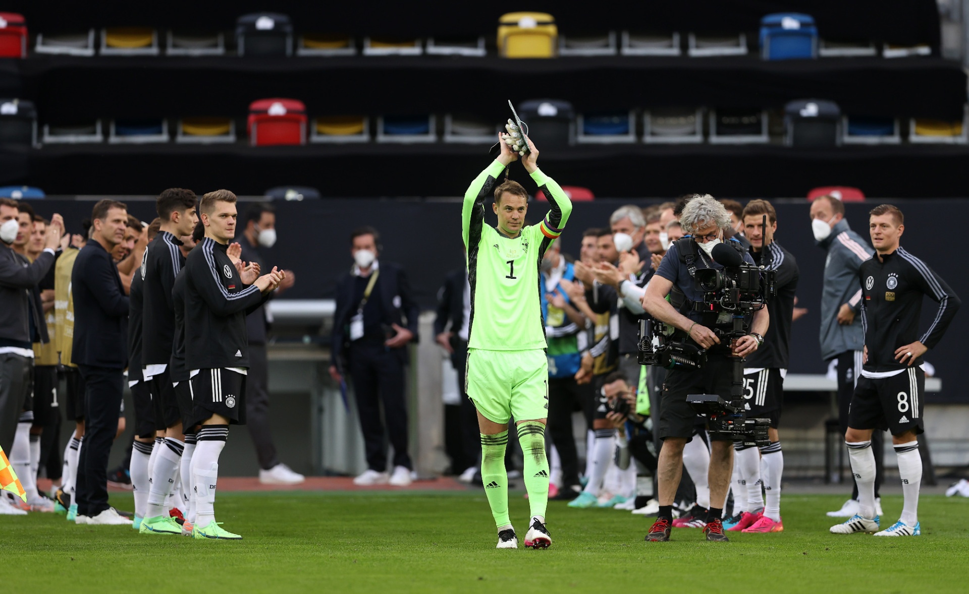[{"clang_id":1,"value":"Fußballspieler Manuel Neuer, im Trikot mit Nummer 1, wird von Teamkollegen und Betreuern nach einem Spiel applaudiert. Hintergrund: leere Zuschauertribünen."}]