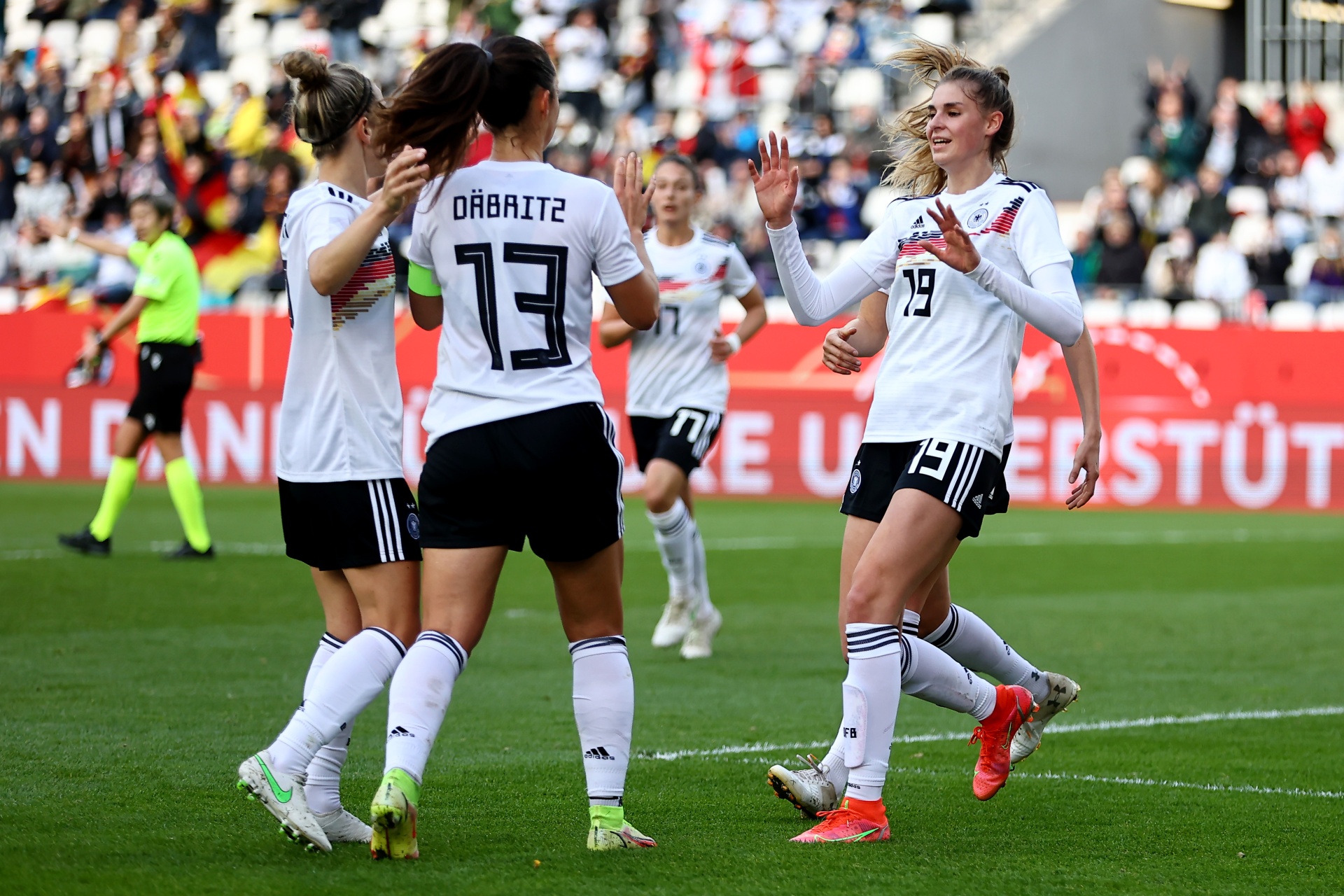 [{"clang_id":1,"value":"Frauenfußballspielerinnen in weißen Trikots mit schwarzen Shorts und roten/blauen Sportschuhen feiern auf grünem Rasen. Im Hintergrund Zuschauer und Werbebanner."}]
