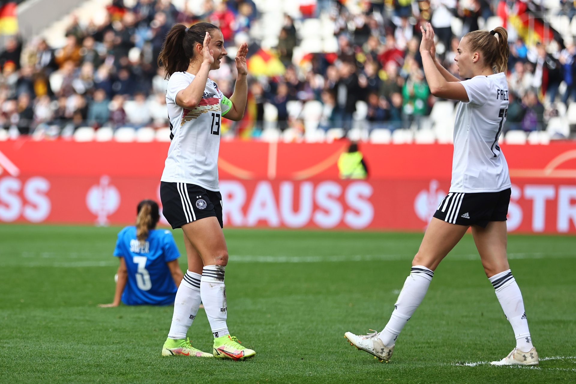 [{"clang_id":1,"value":"Zwei Fußballspielerinnen in schwarz-weißen Trikots der deutschen Nationalmannschaft jubeln auf einem grünen Rasen vor Zuschauern. Im Hintergrund eine Werbetafel."}]