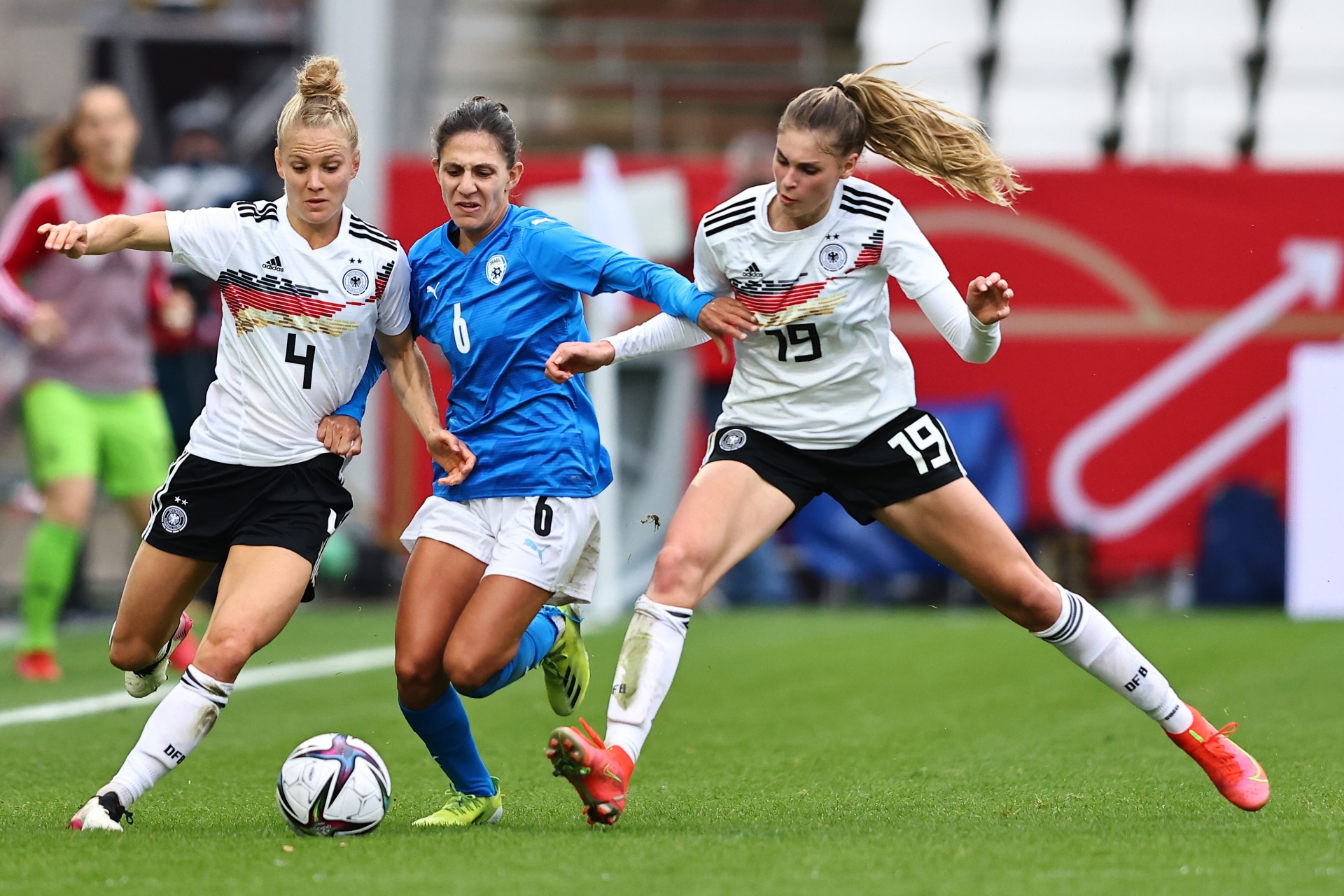 [{"clang_id":1,"value":"Drei Fußballspielerinnen im Wettkampf. Zwei tragen Deutschlandtrikots (Nummern 4 & 19), eine Israel-Trikot (Nummer 6). Fokus auf Ballbesitz und Zweikampf. Rasenplatz."}]