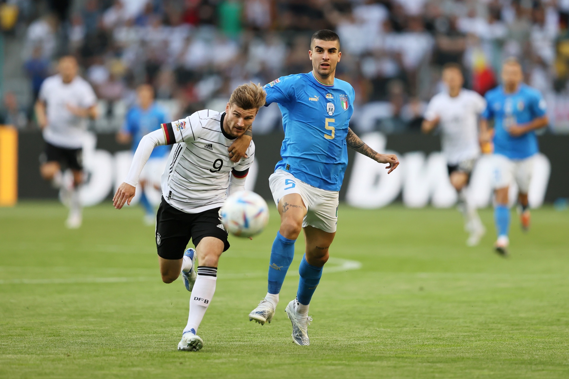 [{"clang_id":1,"value":"Zwei Fußballspieler im Spiel: Links Spieler mit schwarzem Trikot und Nummer 9, rechts Spieler mit blauem Trikot und Nummer 5, beide in Bewegung auf grünem Rasen. Hintergrund mit Zuschauern."}]