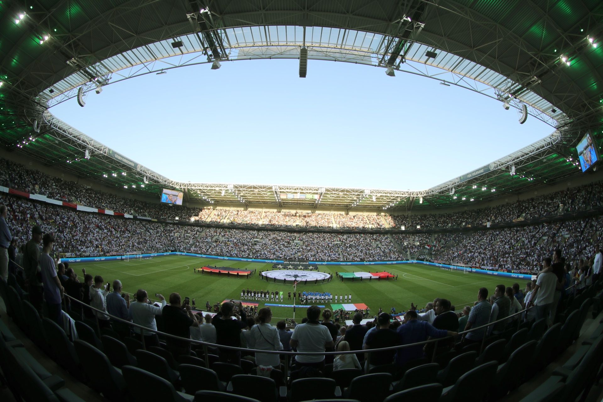 [{"clang_id":1,"value":"Fußballstadion gefüllt mit Zuschauern. Auf dem Rasen eine Zeremonie mit Personen in Kostümen. Blaue und weiße Fahnen sind zu sehen."}]