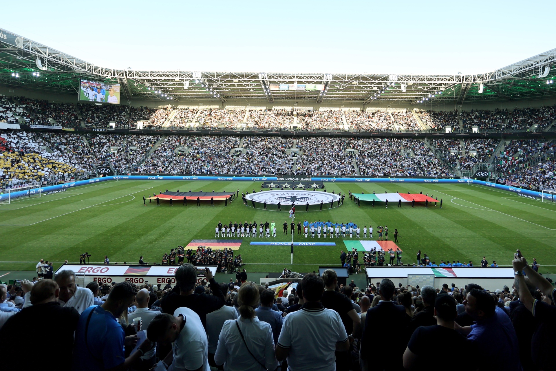 [{"clang_id":1,"value":"Fußballstadion gefüllt mit Zuschauern. Spieler stehen im Kreis auf dem Rasen, umgeben von Fahnen. Werbung für \"ERGO\" ist sichtbar."}]