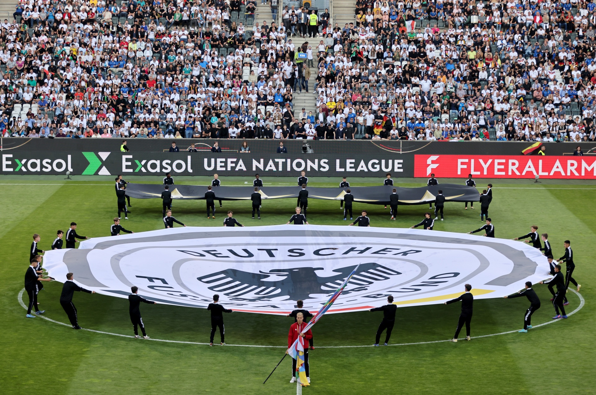 [{"clang_id":1,"value":"Fußballstadion mit vollem Publikum. Spieler stehen um eine große Deutschland-Flagge auf dem Rasen, umgeben von schwarz gekleideten Personen. Werbung für Exasol und FlyerAlarm."}]