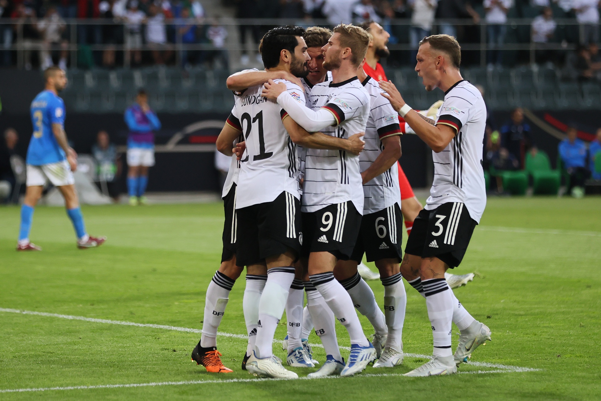 [{"clang_id":1,"value":"Fußballspieler in weißen Trikots feiern ein Tor. Im Hintergrund sind Zuschauer und ein Spieler mit blauem Trikot zu sehen. Rasenfläche im Vordergrund."}]