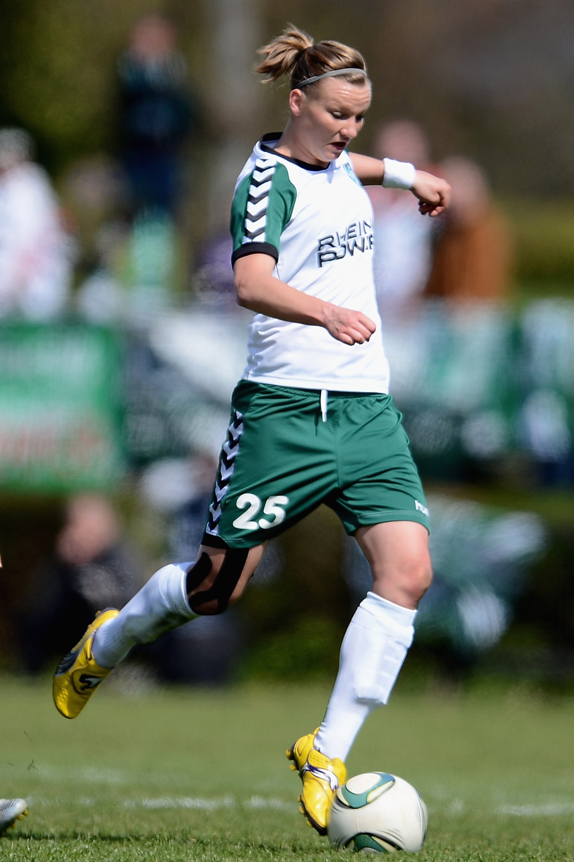 [{"clang_id":1,"value":"Fußballspielerin mit blondem Pferdeschwanz in grün-weißem Trikot und Shorts dribbelt einen Ball auf einem Rasenplatz. Rückennummer 25. Zuschauer im Hintergrund."}]