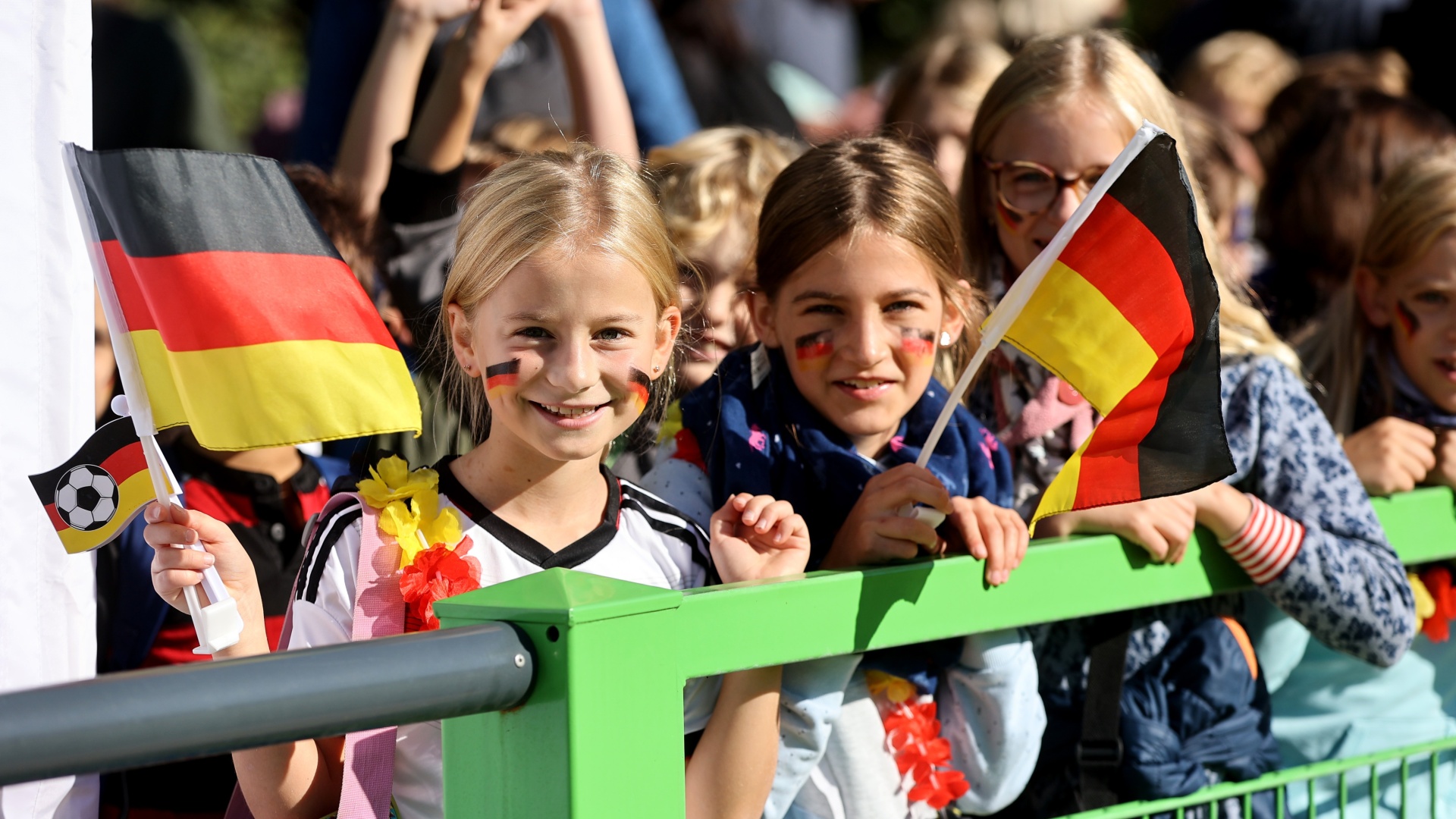 [{"clang_id":1,"value":"Kinder mit bemalten Gesichtern und Deutschlandflaggen sitzen auf einer Tribüne. Im Hintergrund weitere Zuschauer und ein Fußball."}]