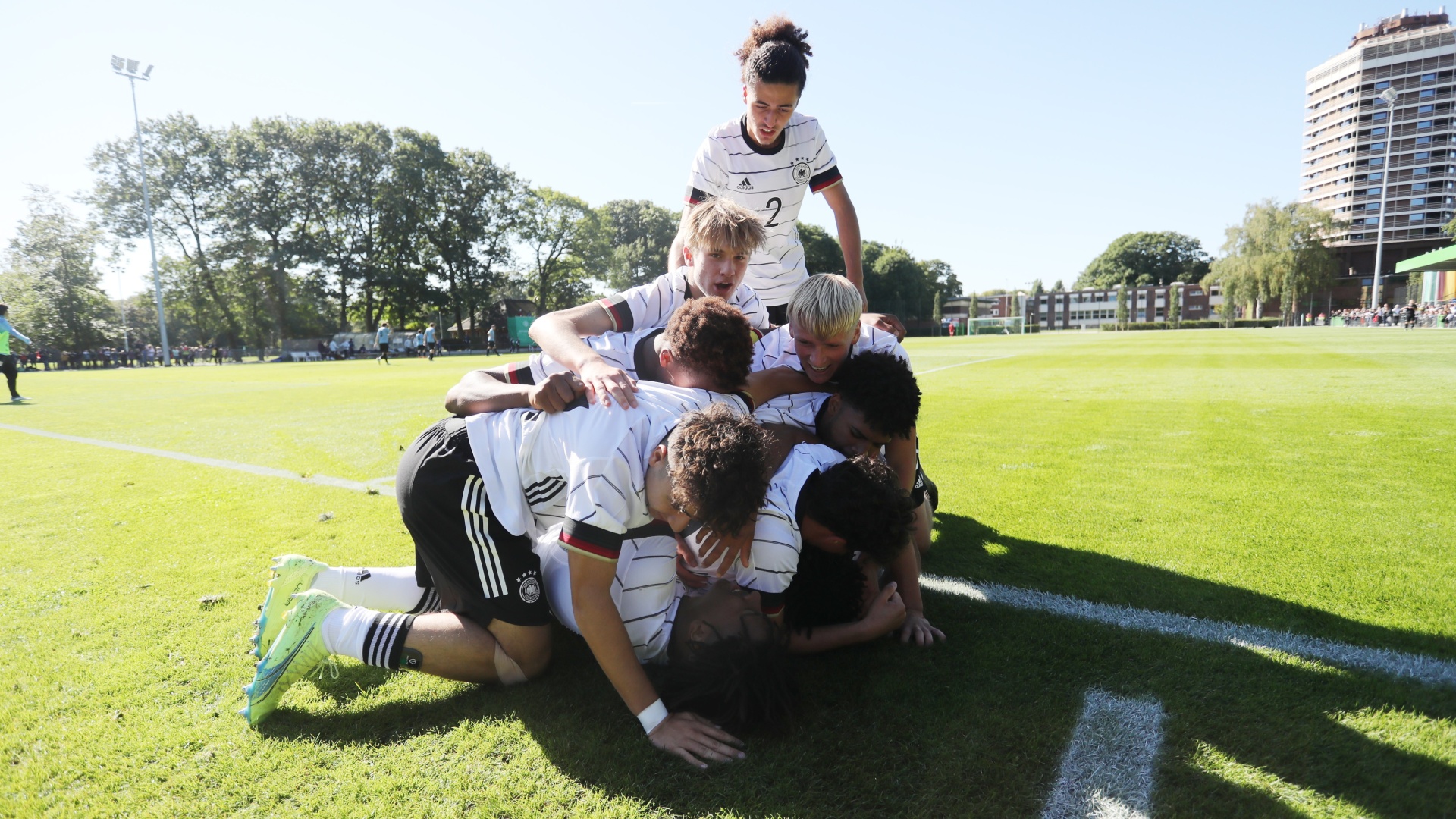 [{"clang_id":1,"value":"Fußballspieler in Deutschlandtrikots feiern einen Treffer im Grünen. Mehrere Spieler liegen übereinander, ein weiterer steht und jubelt. Im Hintergrund Gebäude und Bäume."}]