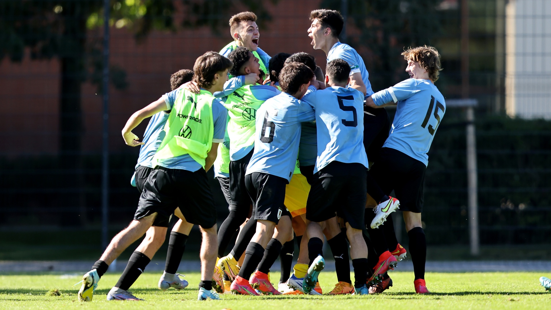 [{"clang_id":1,"value":"Fußballspieler in blauen und schwarzen Trikots feiern einen Treffer eng umschlungen auf dem Rasen. Einige tragen gelbe Westen. Im Hintergrund Bäume und ein Zaun."}]