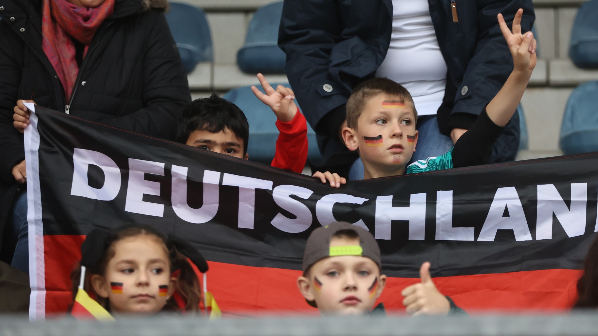 [{"clang_id":1,"value":"Zuschauer bei Sportveranstaltung. Vier Kinder mit Deutschlandflaggen-Gesichtsbemalung und -Banner. Kinder heben Hände. Erwachsene im Hintergrund."}]