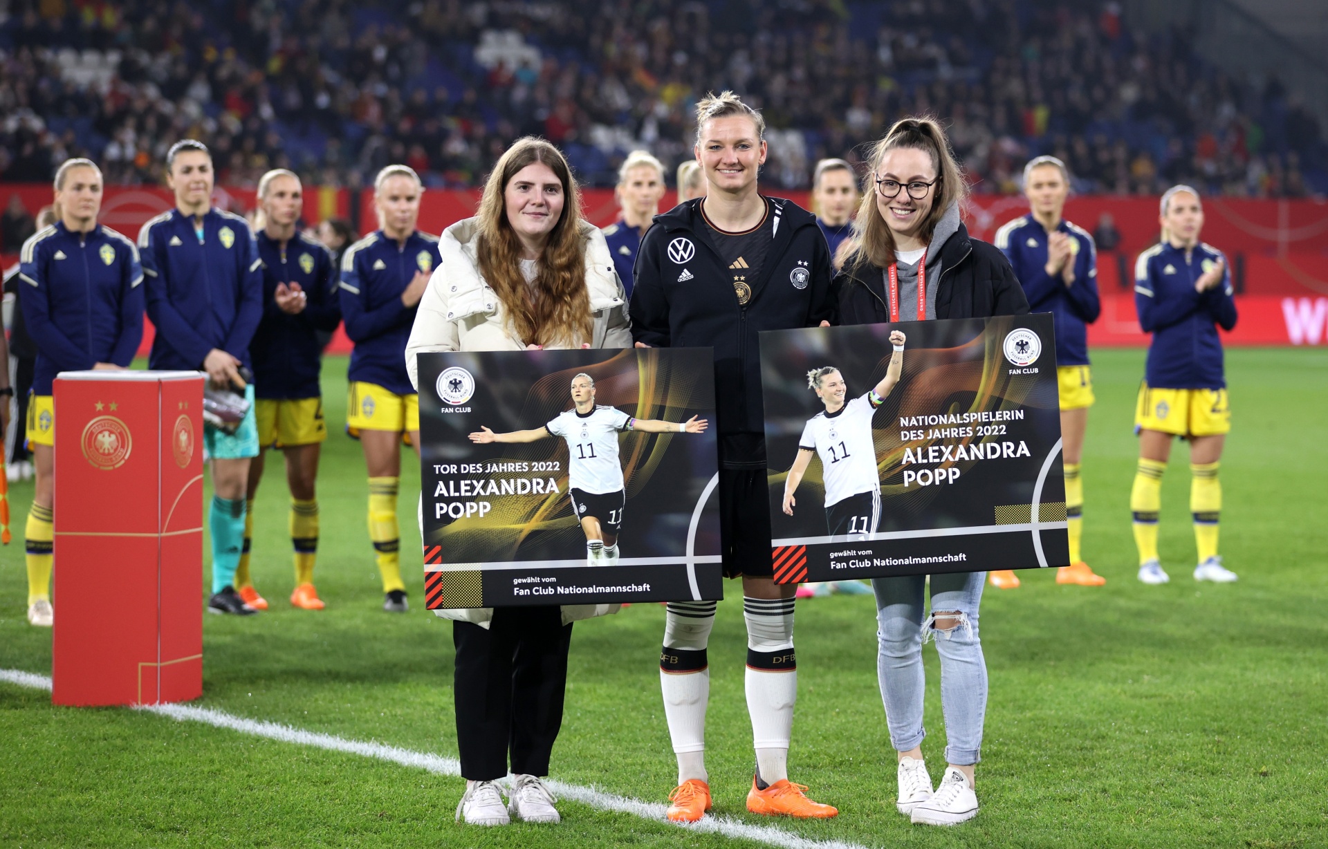 [{"clang_id":1,"value":"Alexandra Popp, Fußballspielerin, mittig mit zwei Auszeichnungen für \"Tor des Jahres\" und \"Nationalspielerin des Jahres\" auf einem Fußballfeld stehend. Im Hintergrund Fußballspielerinnen."}]