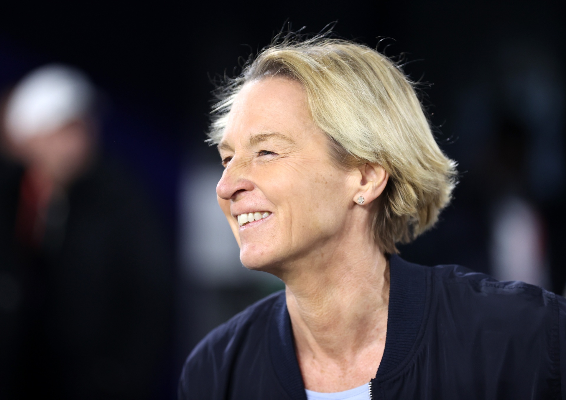 [{"clang_id":1,"value":"Frau mit blonden, kurzgeschorenen Haaren lächelt in die Kamera. Sie trägt eine dunkle Jacke. Im Hintergrund verschwommene Personen und ein Sportfeld."}]