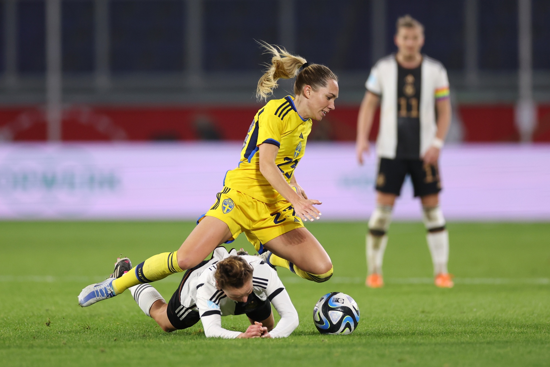 [{"clang_id":1,"value":"Zwei Fußballspielerinnen kämpfen um den Ball auf grünem Rasen. Eine Spielerin in gelbem Trikot springt über eine gefallene Spielerin in schwarzem Trikot. Im Hintergrund eine weitere Spielerin."}]